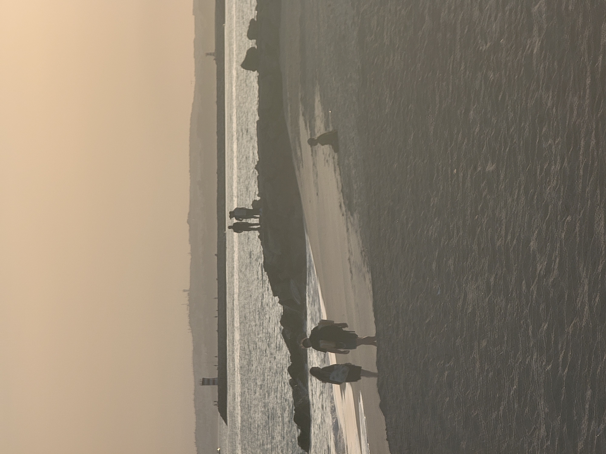 The image depicts a serene beach scene at dusk in Quarteira, Portugal. The sandy shore is sparsely populated. In the foreground, two individuals are walking on the wet sand near the water's edge, one carrying a tote bag. Further back on the beach, a solitary figure sits on the sand. In the middle ground, a group of people are silhouetted against the sea, standing on a rocky breakwater. The breakwater extends into the ocean and is punctuated by a lighthouse. The sea itself is calm, reflecting the soft, hazy light of the setting sun, which casts a warm, golden hue over the entire scene. The horizon is marked by a distant coastline with subtle hints of structures and a second lighthouse. The overall atmosphere is peaceful and contemplative.