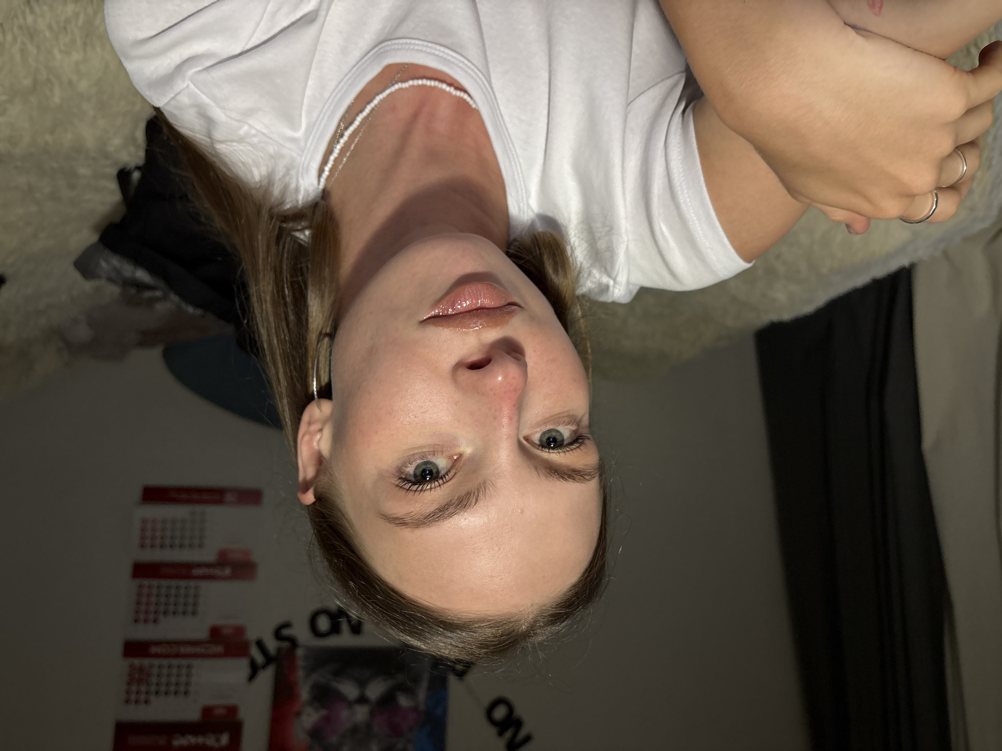The image depicts a young adult female from a chest-up perspective, posed for a selfie indoors in what is identified as a location within the city of Perm, Russia. She has light skin, light brown hair parted centrally, and light blue-green eyes. Her lips are slightly parted and covered with a glossy, light brown product. She wears a white crew-neck t-shirt, two thin silver rings on her left ring finger, and layered white beaded necklaces. A small silver hoop earring is visible in her right ear. Her left arm is bent, with her hand resting on her forearm.
The background shows a light-colored wall with several decorations. To the far left, a dark curtain is partially visible. Above her head, partially obscured black letters spell "NO ST" (likely part of a larger phrase). A poster depicting a stylized cat face with pink accents is partially visible behind her upper right head. Further to the right are vertically stacked red rectangular items resembling calendar pages with white grid patterns and text. The woman appears to be seated on a light-colored, fluffy textile surface, possibly a bed or rug. A dark bag and a dark blue pillow are partially visible in the lower right background.