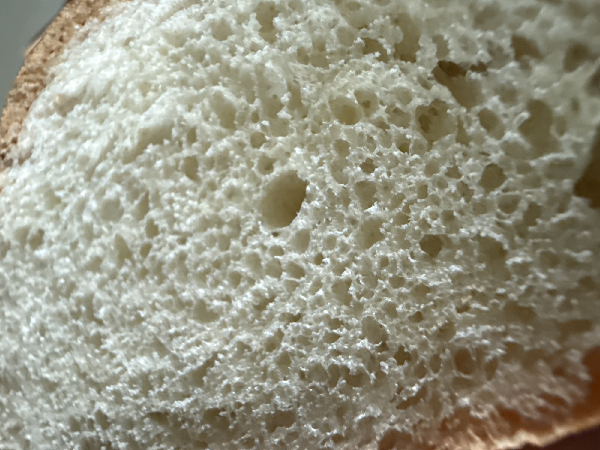 The image is a close-up, macro photograph of a slice of white bread. The texture of the bread's crumb is visible, showing a porous structure with many small air bubbles. The crust of the bread is visible along the left and top edges of the image, appearing golden brown. The lighting creates shadows within the pores, highlighting the uneven surface. There are no people, objects, actions, interactions, or environmental details present in this image that would allow for identification of a location in Okhtyrka, Ukraine.