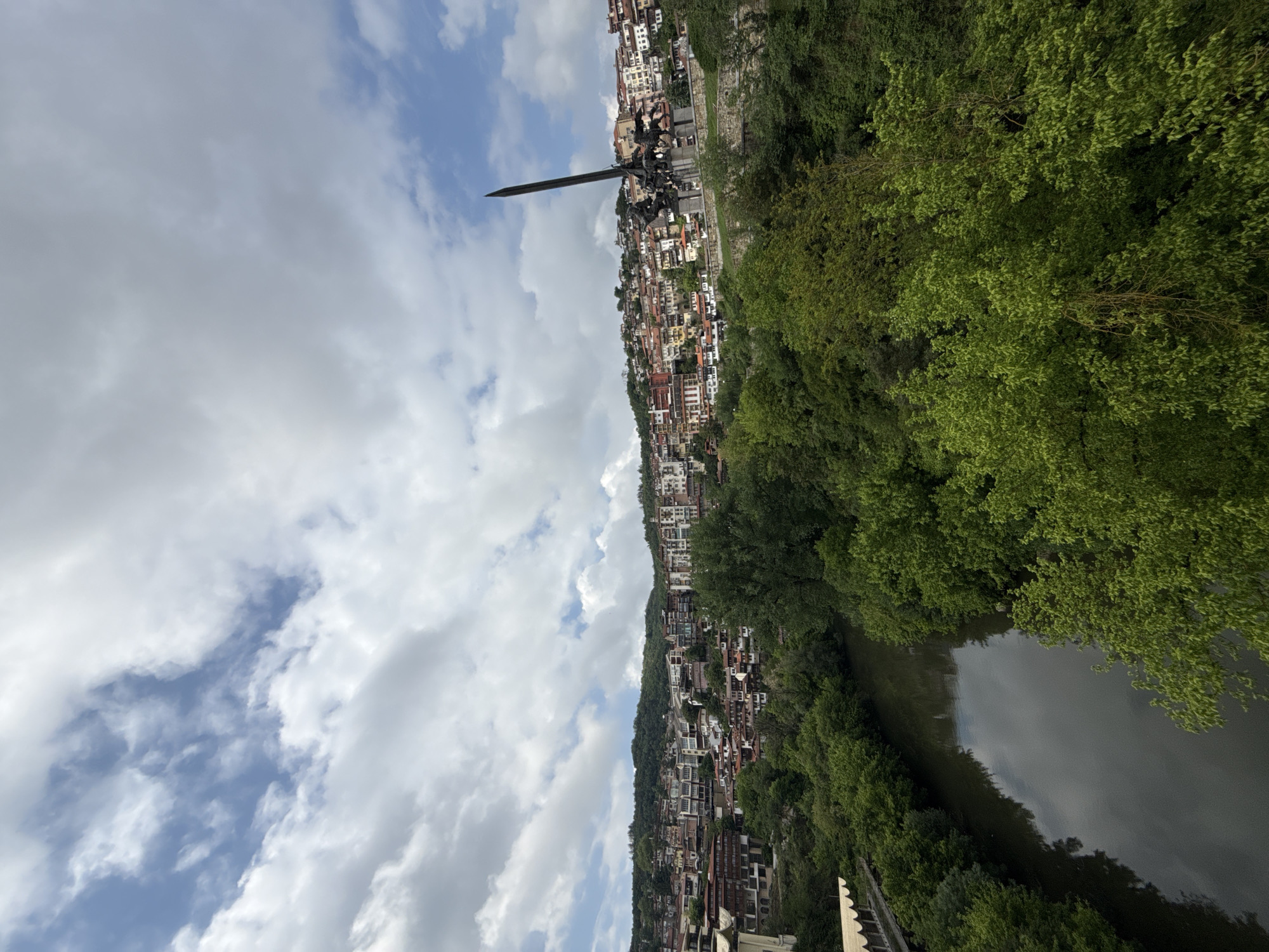 The image is an outdoor, landscape shot of Veliko Tarnovo, Bulgaria. The camera angle is at a low-level looking up at a hillside filled with multi-story, residential buildings with red rooftops. The sky is partially cloudy. A river runs along the bottom-right side of the image, separated from the hillside by a thick line of trees. A large black, metal sword is erected as part of a statue in the city.