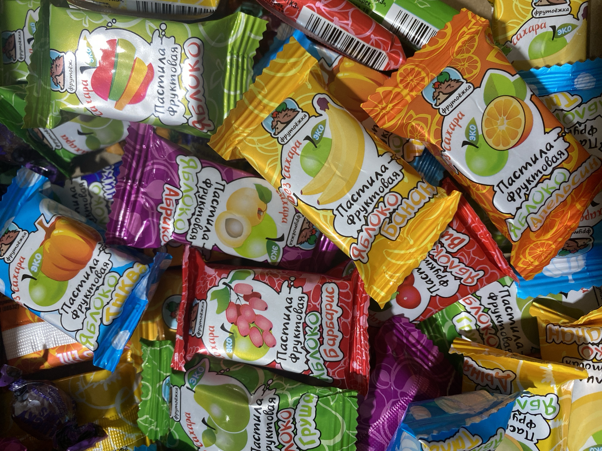 A close-up image shows a vibrant assortment of individually wrapped fruit pastilles, piled together. The wrappers are brightly colored and feature illustrations of various fruits along with Cyrillic text. Each snack belongs to the "Фрутоёжка" (Frutozhka) brand, identified by a small hedgehog logo.
Common textual elements visible on the packaging include "эко" (eco), "без сахара" (sugar-free), and "Пастила Фруктовая" (Fruit Pastille). Numerous flavors are identifiable:
* **Green wrappers** display "ЯБЛОКО" (Apple) with a sliced apple and "ЯБЛОКО ГРУША" (Apple Pear) with a pear.
* **Yellow wrappers** show "ЯБЛОКО БАНАН" (Apple Banana) with bananas and an apple.
* **Orange wrappers** feature "ЯБЛОКО АПЕЛЬСИН" (Apple Orange) with an orange slice and an apple, and "ЯБЛОКО ТЫКВА" (Apple Pumpkin) with a pumpkin.
* **Red wrappers** highlight "ЯБЛОКО БАРБАРИС" (Apple Barberry) with barberries and "ЯБЛОКО ВИШНЯ" (Apple Cherry) with cherries.
* **Purple wrappers** illustrate "ЯБЛОКО АБРИКОС" (Apple Apricot) with apricots.
The wrappers are glossy and appear slightly crinkled, suggesting soft contents. Bright lighting creates reflections on the shiny surfaces, enhancing the colorful presentation. The overall impression is of a wholesome, fruit-based snack assortment. This scene is set in Verkhnevesyoloye, Russia.