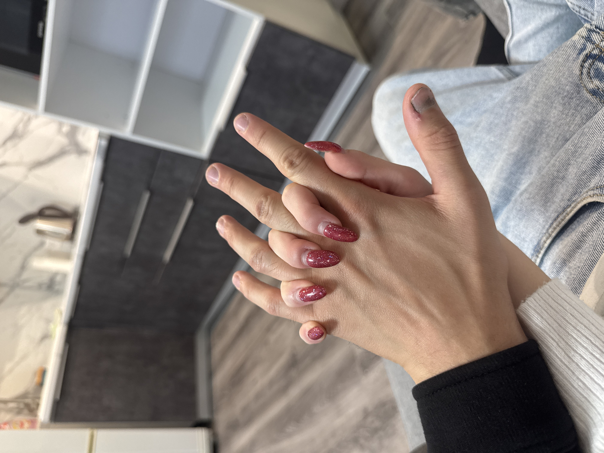 Two people are holding hands in a domestic setting. The person on the left is wearing a black sweatshirt and the person on the right is wearing a white long-sleeved shirt and blue jeans. The person on the left has nails painted with sparkly red polish. The person on the right has a bruised thumb. In the background, a kitchen counter with cabinets and a white shelf unit are visible. The floor appears to be wooden laminate.
