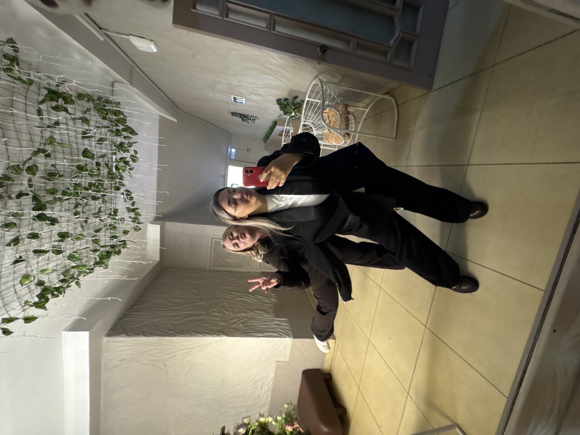 Two women are posing for a selfie in an indoor location with tiled floors. The first woman is holding a red smartphone and wearing glasses, a white shirt, and a black blazer with matching pants. The second woman, beside her, is wearing a dark jacket and pants, and white sneakers, and is making a peace sign with her hand.
The background includes a white metal chair, a door with glass panels, and a wall decorated with greenery and lights. There is also a brown object and flowers visible at the bottom of the frame.
There is no location information present in the image.