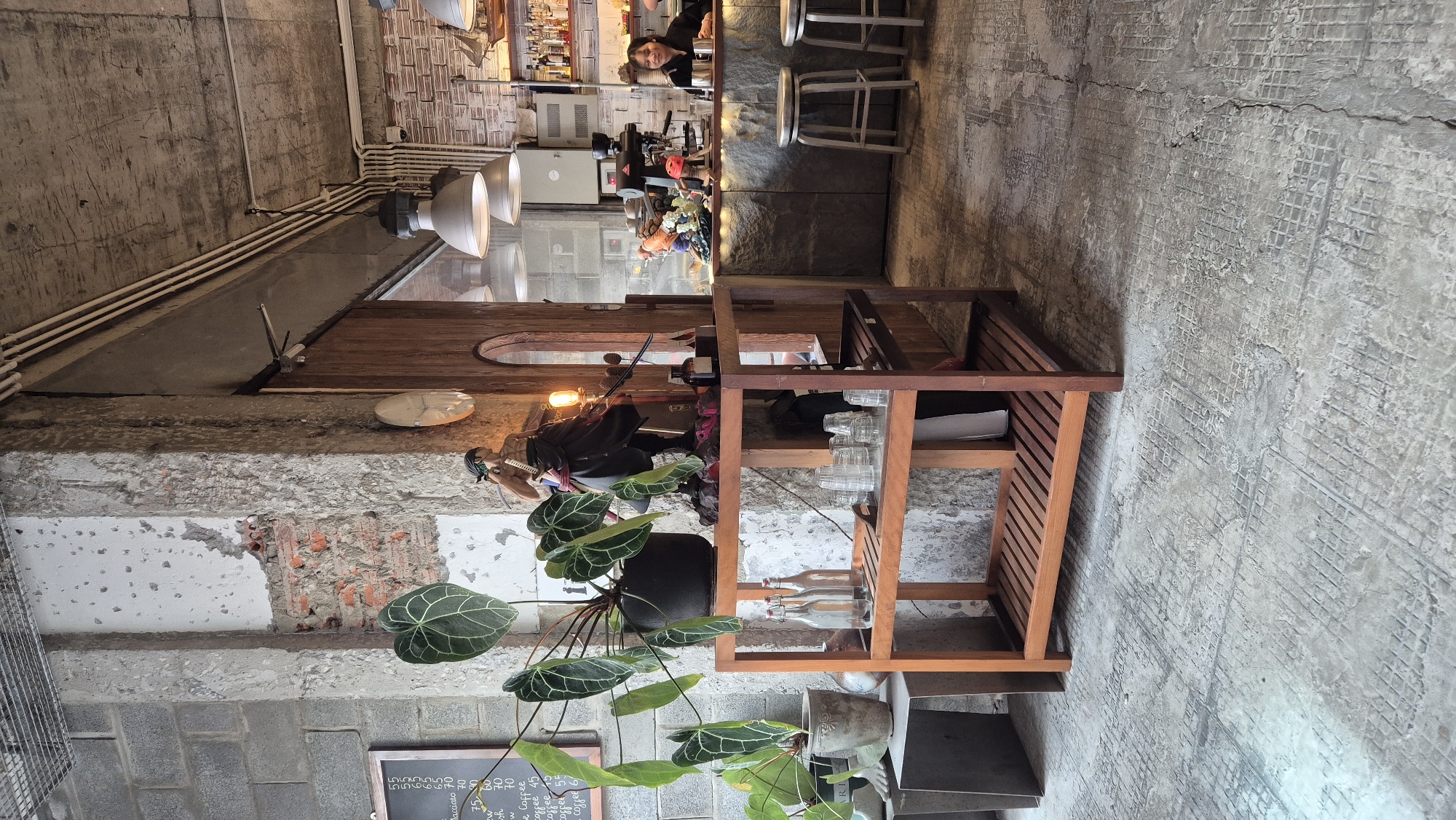 The image shows the interior of a cafe with a raw, industrial aesthetic in Đà Nẵng, Vietnam. Exposed brick and concrete walls, along with a concrete floor, create a textured and unfinished look. A wooden shelf unit stands in the foreground, holding decorative items, including a figurine of a character from One Piece, a large potted plant with dark green leaves, and several glass bottles and drinking glasses.
Behind the shelf, a wooden counter area is visible, where a barista is working. A coffee machine is present, and shelves behind the counter are stocked with bottles and glassware. Two patrons are seated at the counter, with one woman in a black shirt visible. Hanging industrial-style pendant lights illuminate the space. The overall atmosphere suggests a trendy and modern cafe environment.