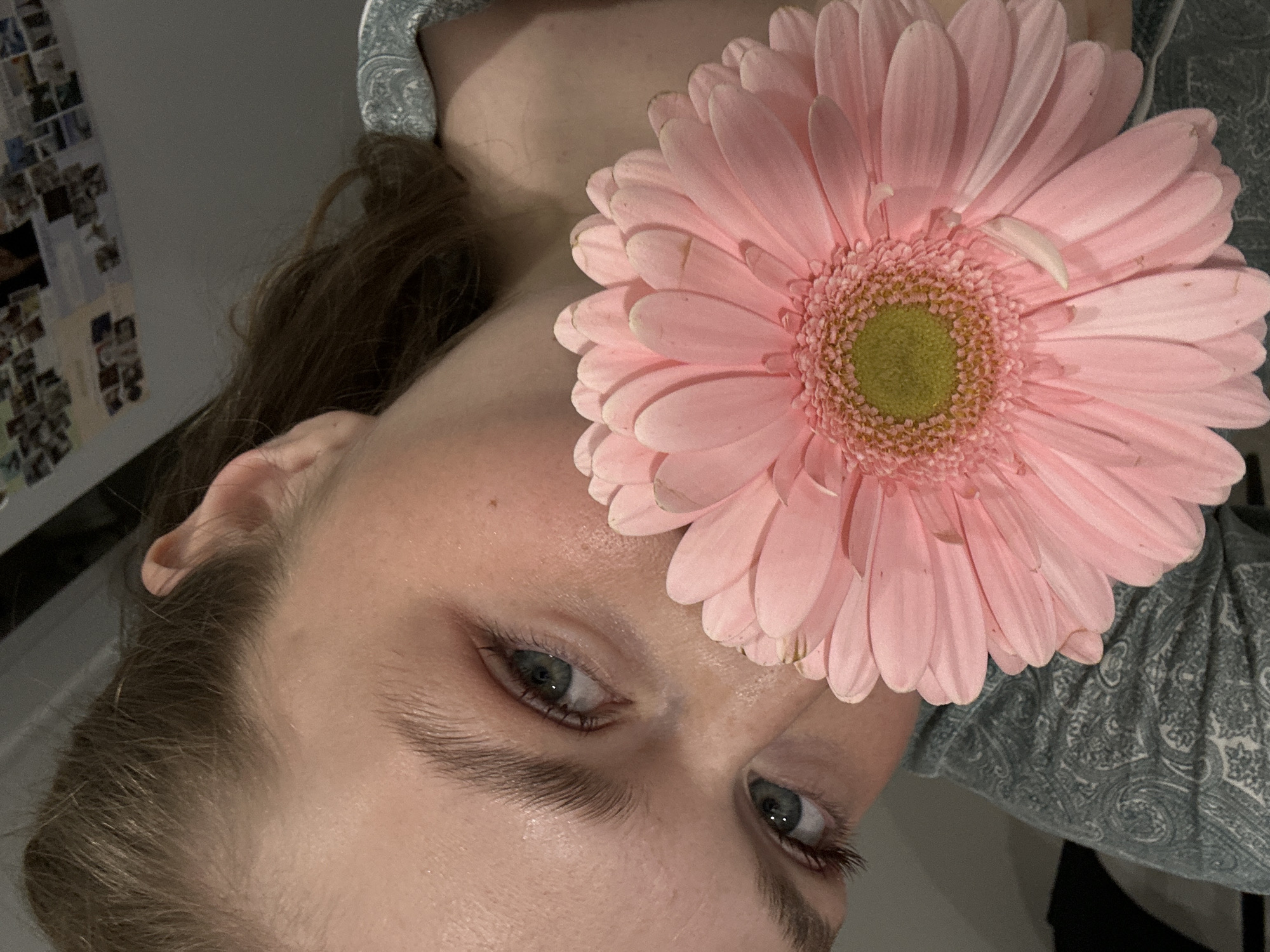 A close-up, tilted portrait shows a light-skinned person with brown hair. A large pink gerbera daisy flower covers the center of their forehead. The person has light blue eyes and defined eyebrows. The background includes a wall covered with what appear to be photos, and a paisley patterned fabric around the neck. The location is not evident from this image and cannot be determined to be Kazan, Russia.
