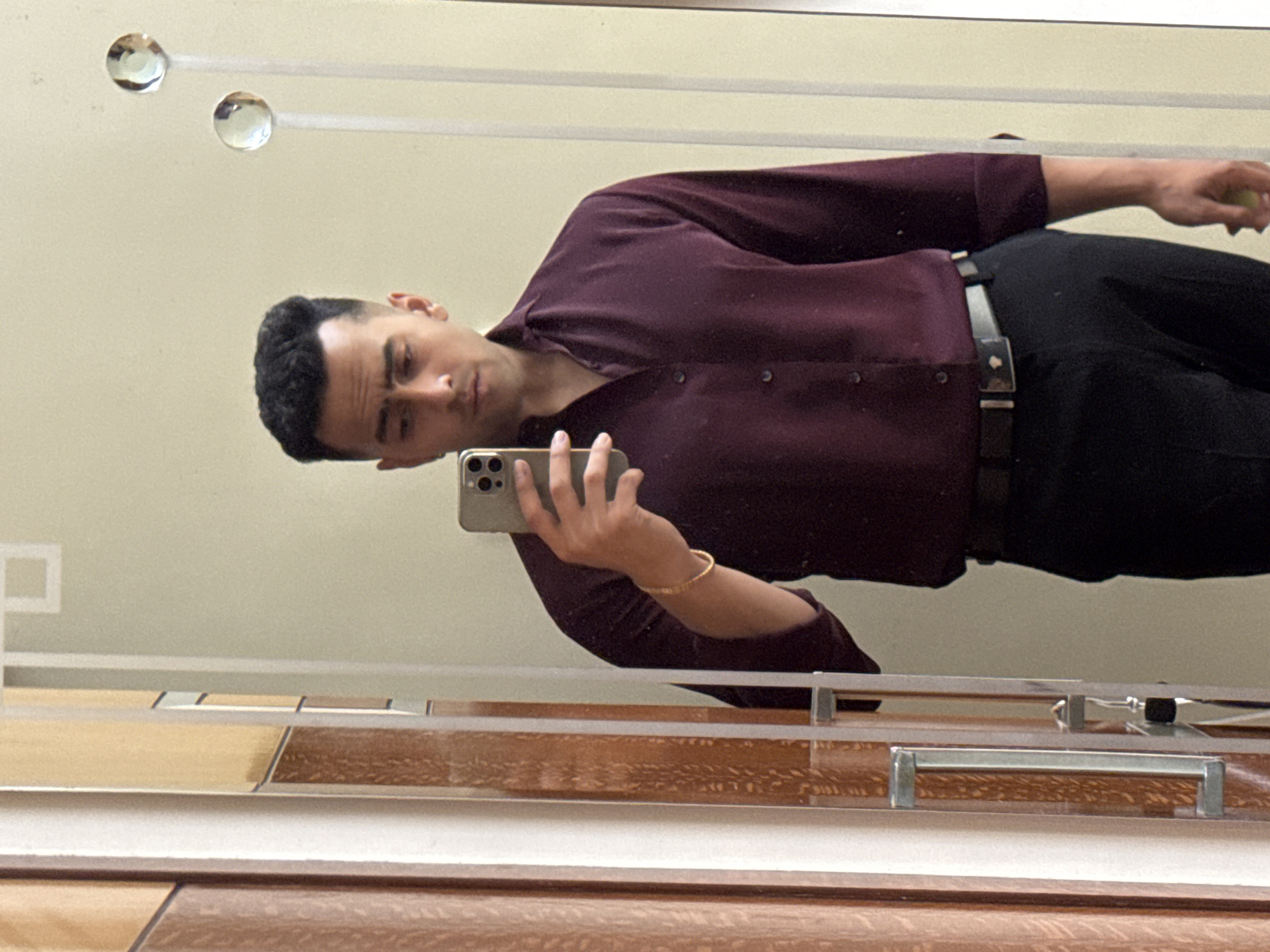A man is reflected in a mirror while taking a selfie with a smartphone. He is wearing a burgundy button-down shirt and black pants with a black belt. The mirror has decorative horizontal lines and circular embellishments at the top. The bottom of the mirror reflects a wooden surface with metal handles, possibly part of a cabinet or vanity. The location is unknown and there is no mention of Kolkata, India.