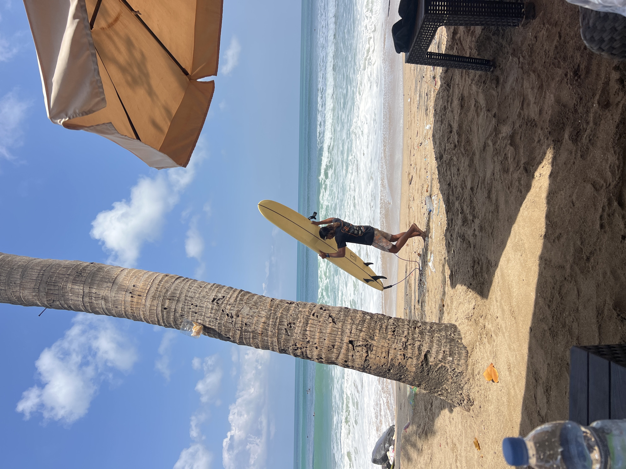 A person is holding a yellow surfboard and walking on the beach in Kuta, Indonesia. The person is wearing a dark t-shirt and patterned shorts. A palm tree is to the left of the person, and a beach umbrella is to the right. The ocean is in the background with waves breaking. There are a few other surfers in the water. The beach is sandy with some debris.