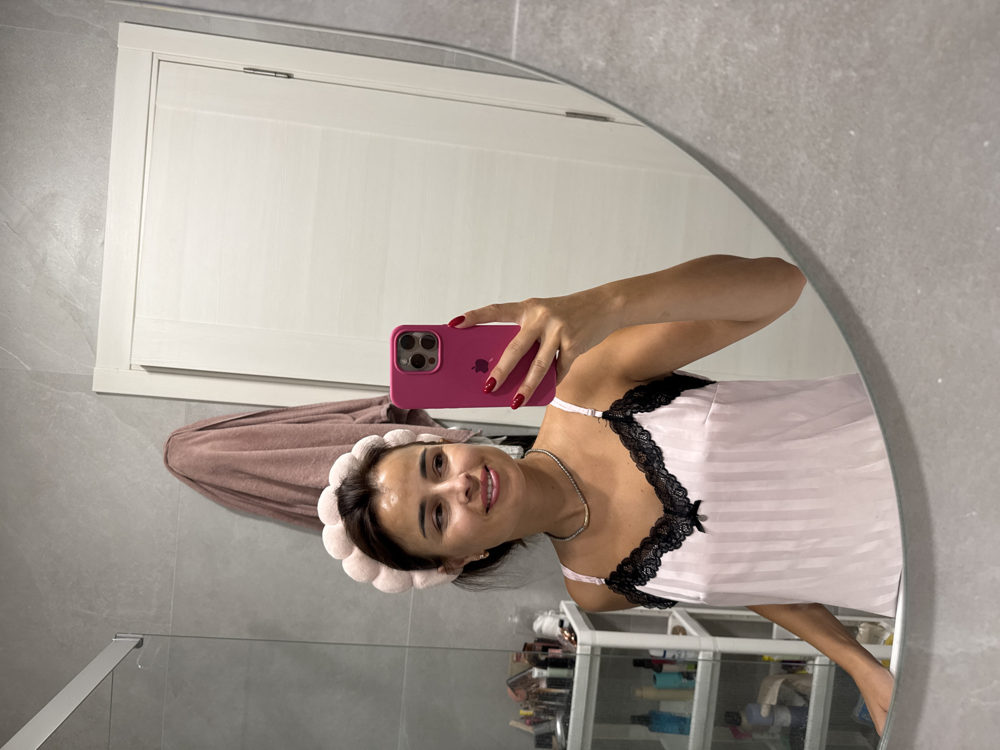 A woman with brown hair and a pink headband is taking a selfie in a bathroom mirror. She is wearing a pink striped slip with black lace trim and a small bow. She is also wearing a silver necklace and has red nail polish. In the background, there is a white door, a towel hanging on the wall, and shelves filled with toiletries. The bathroom has grey tiled walls.