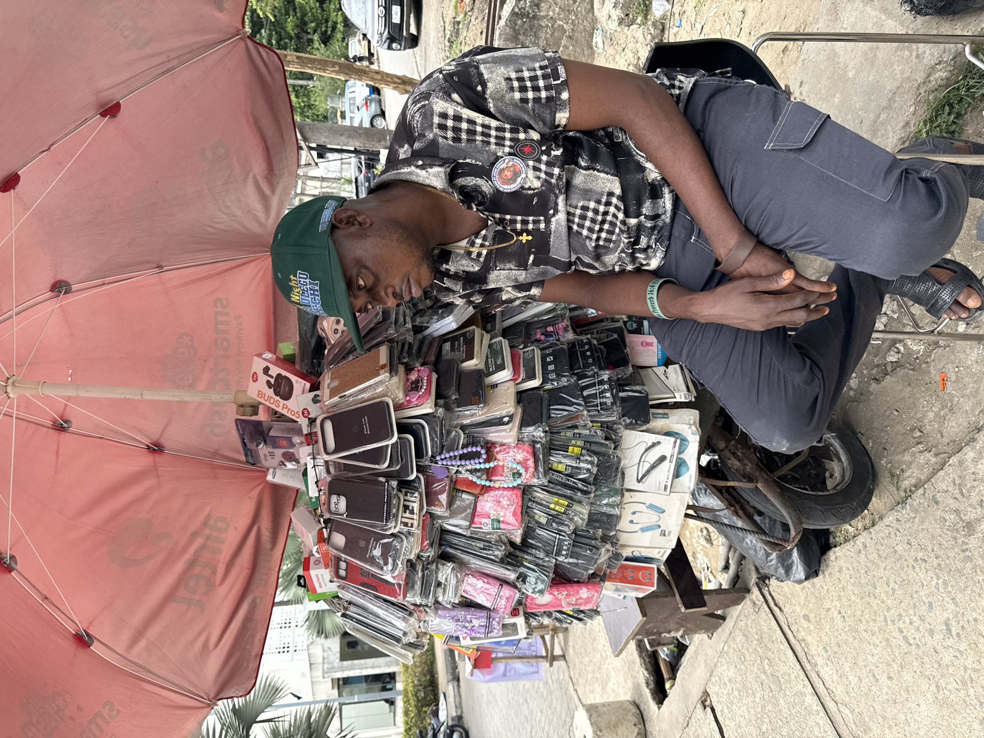 In Itirin, Nigeria, an African man wearing a green baseball cap, patterned short-sleeve shirt, and gray pants sits on a chair behind a makeshift stand covered with packaged cell phone accessories. The stand is under a faded red umbrella. The man appears to be dozing, his head resting on the merchandise. The stand is a bicycle wheel covered with gray plastic.