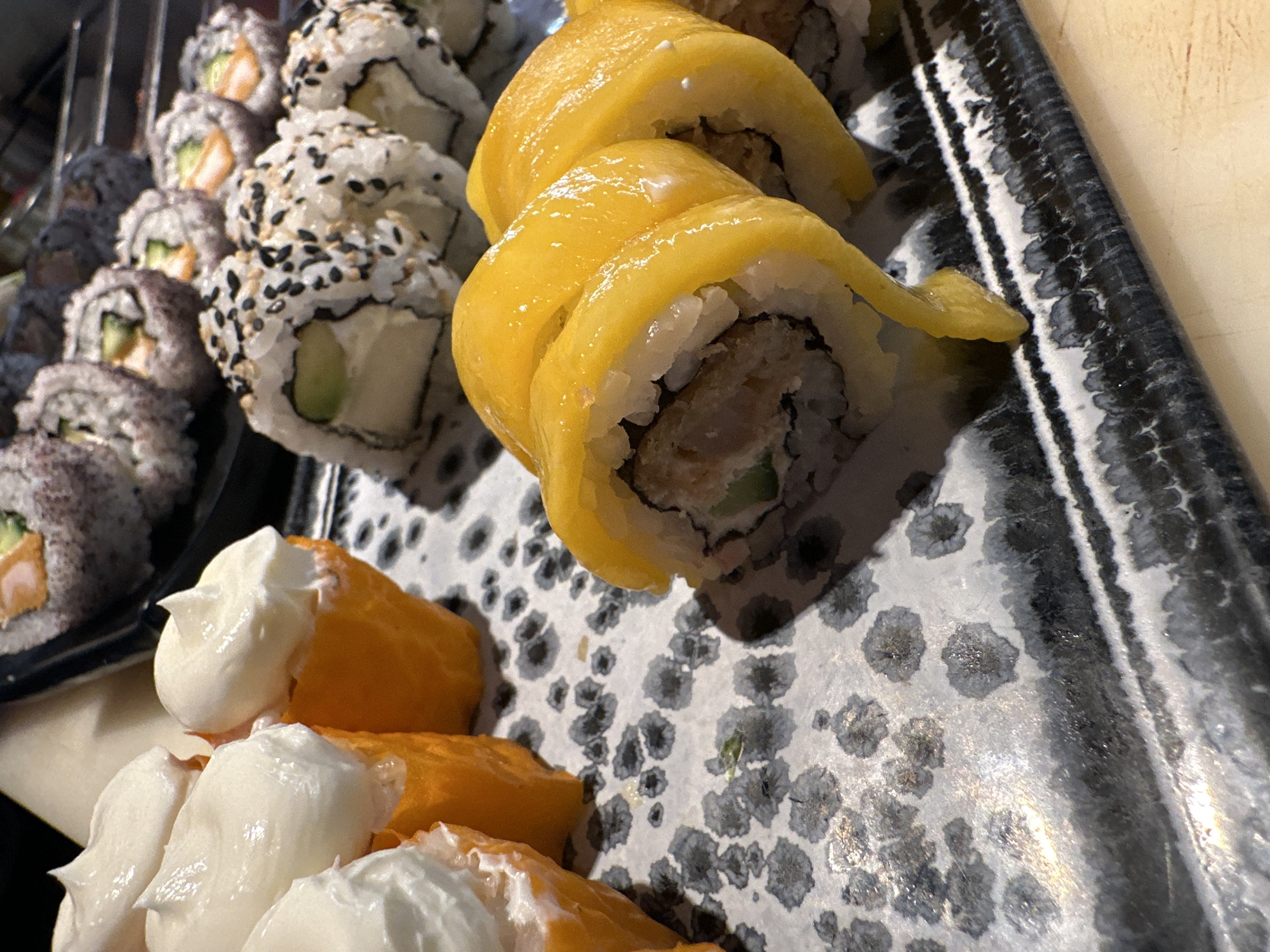 The image shows an assortment of sushi on a patterned tray. There are several types of sushi including rolls covered in mango slices, rolls with sesame seeds, and rolls with tempura batter. Pieces of what appears to be salmon nigiri topped with mayonnaise are also visible. The scene is not identifiable as being in Sofia, Bulgaria.