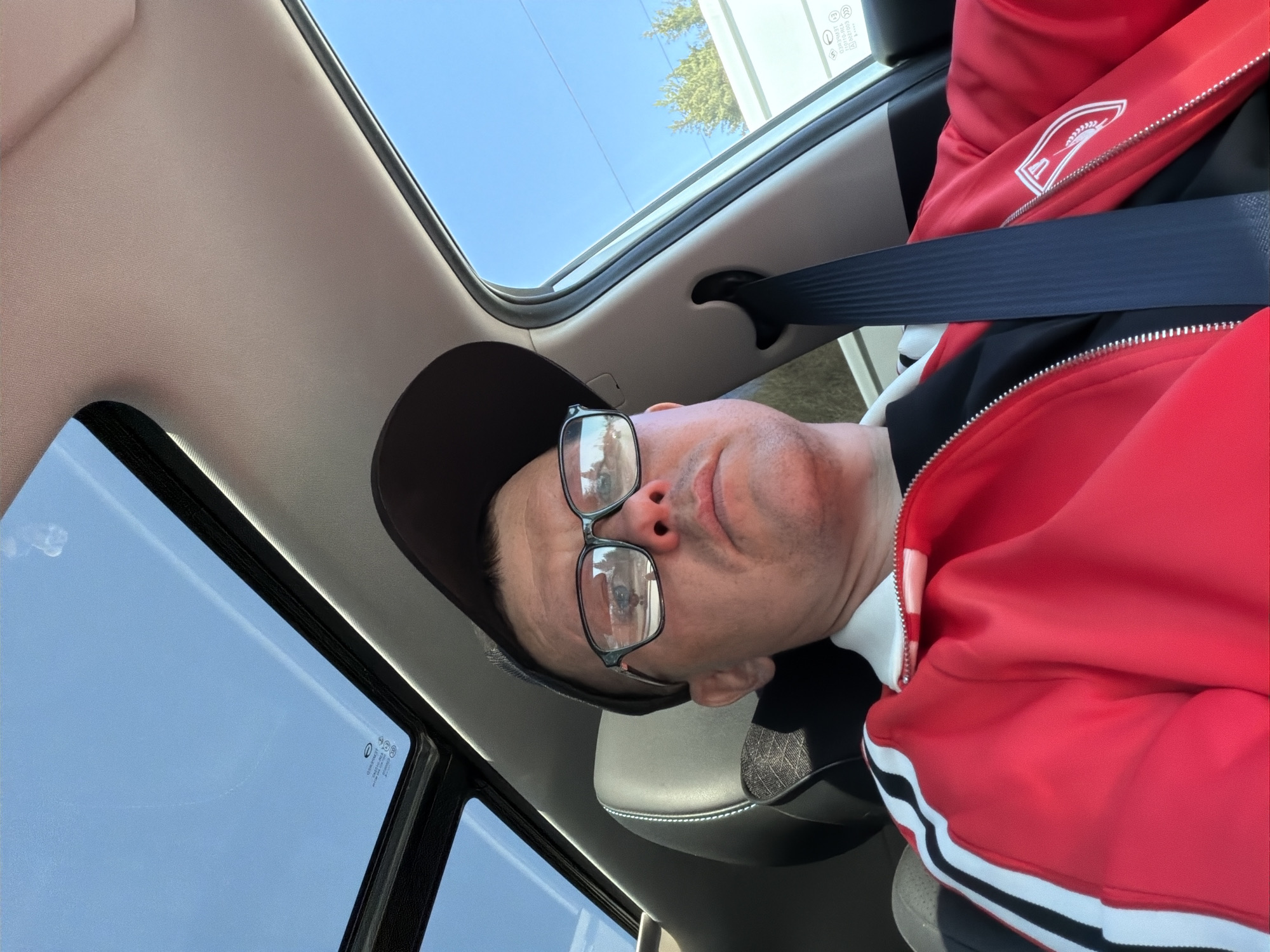 The image shows a man wearing a black cap and glasses in the passenger seat of a car. He is wearing a red jacket with white stripes and a black undershirt, and a seatbelt is fastened across his chest. The car has a sunroof and is likely in Chaikovsky, Russia, based on the registration sticker on the window. The view through the sunroof includes a clear sky. The car's interior includes a light-colored headliner.