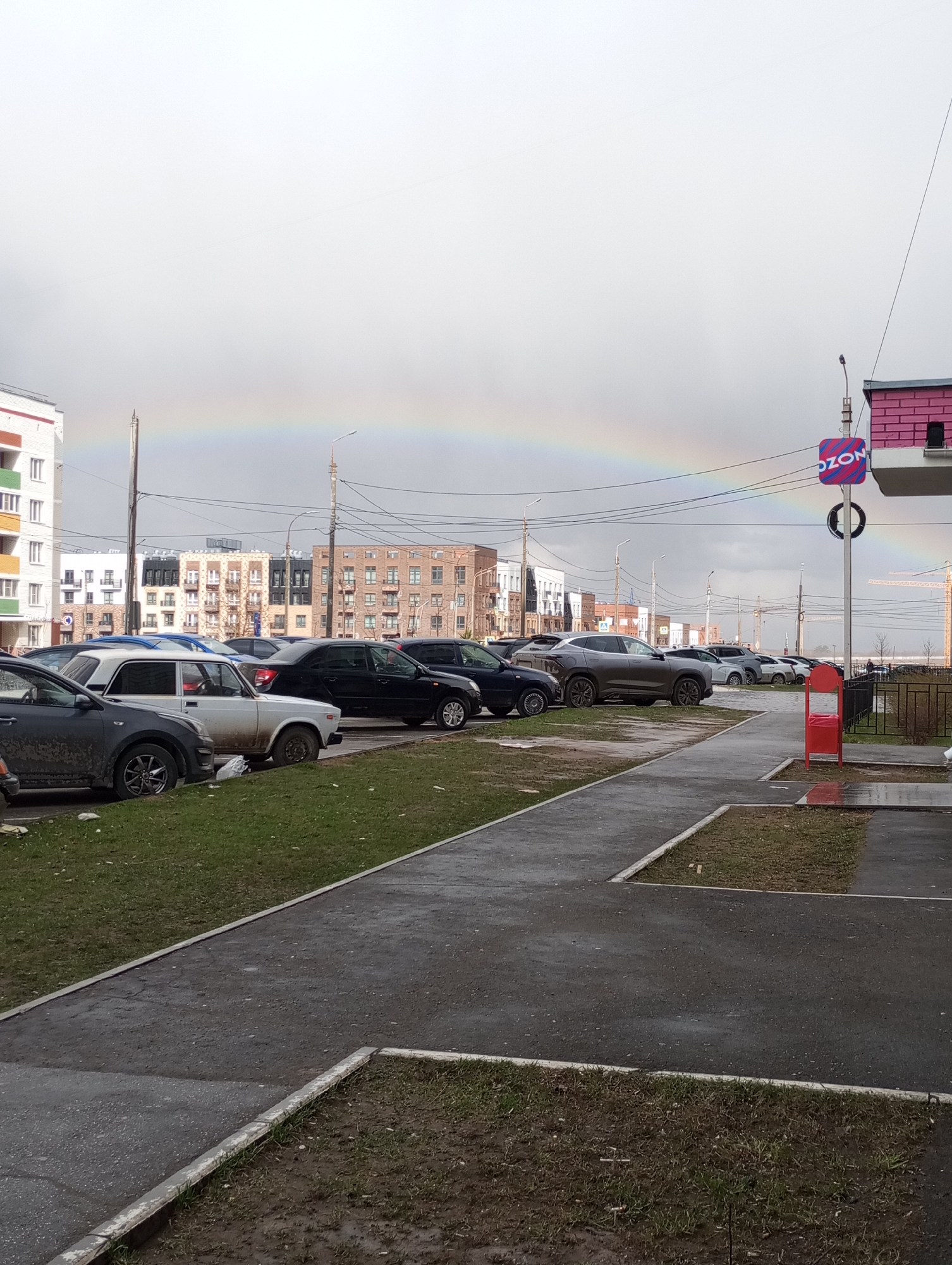 This image depicts an urban street scene in Izhevsk, Russia, characterized by an overcast sky and a prominent rainbow arcing across the upper portion. The ground, including both paved areas and grassy strips, appears wet and muddy, indicative of recent rainfall.
In the foreground, a paved sidewalk borders a patch of muddy grass. To the left, numerous cars are parked along the street and in an unpaved area, many of them visibly dirty. A white, older model sedan with mud splatters and rust is particularly noticeable among the parked vehicles, which include various other sedans and SUVs.
The background is dominated by multi-story residential buildings, showcasing different architectural styles and colors such as white, brown, and beige. Utility poles, laden with electrical wires, are scattered throughout the scene, and streetlights are attached to some of them. A construction crane is faintly visible in the far distance on the right.
On the right side of the frame, a vertical pole supports a blue rectangular sign that clearly displays the word "OZON" in white letters with red and white stripes. Below this, a black circular object resembling a tire or ring is also attached to the pole. A bright red, open-top trash bin stands near a low black metal fence on the sidewalk.
The scene suggests daytime, possibly late afternoon, given the appearance of the rainbow. The weather is transitioning from rain to clearer conditions. No people are visible, and no specific activities are taking place.