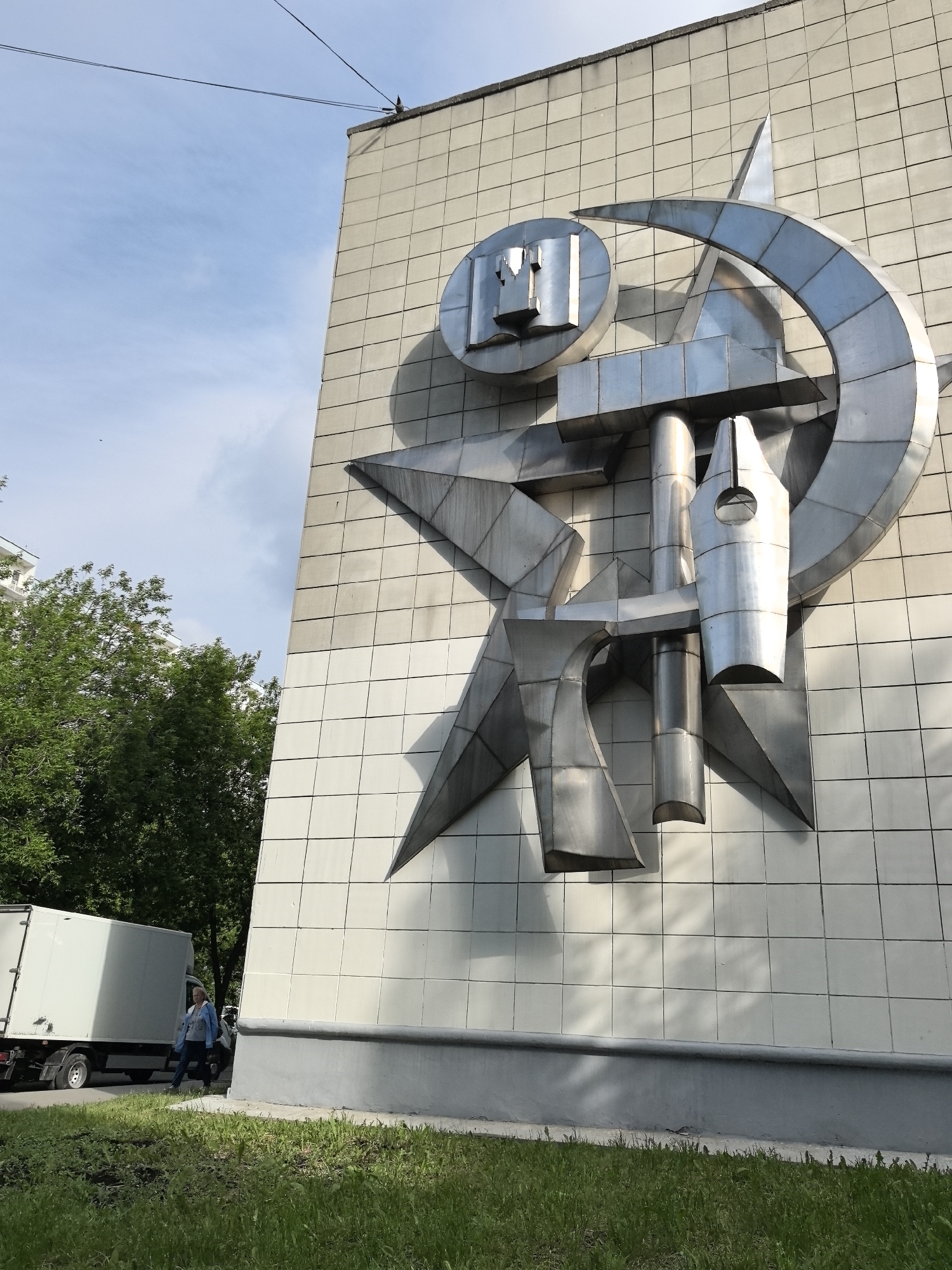 A low-angle, eye-level outdoor shot shows the corner of a building in Perm, Russia. The building has a large, silver, abstract metal sculpture affixed to its tiled facade. The sculpture features geometric shapes, including a star and a circular element containing stylized Cyrillic letters.
To the left, a white box truck is parked on a road adjacent to a grassy area. A woman, dressed in jeans and a light blue jacket, walks along the sidewalk near the truck. Trees are visible behind the truck and woman. The sky above is pale blue with scattered clouds. A power line runs across the top portion of the image.