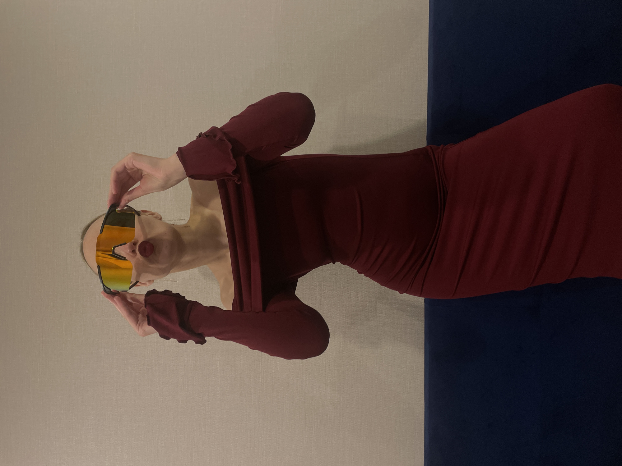 A fair-skinned person with slicked-back hair and long earrings is wearing a dark red, fitted dress with ruffled sleeves and bright yellow sunglasses. The person is holding the sunglasses up to their face. The background consists of a light beige wall on the left and a dark blue surface on the right. The image is rotated counter-clockwise approximately 90 degrees. There is no evidence the image was taken in the city of Подшивалово, Russia.