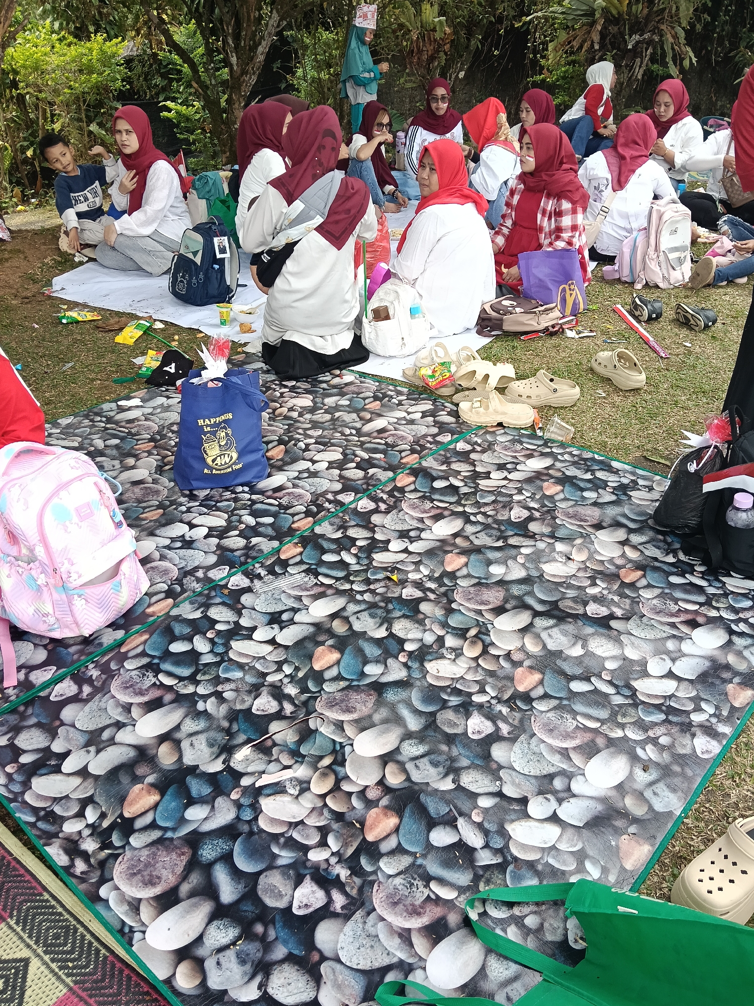 In Cisarua, Indonesia, a group of people are picnicking on a grassy area beneath trees. The foreground is dominated by a large picnic blanket with a stone pattern, upon which a pink backpack with unicorn decals and a blue tote bag with "HAPPINESS IS... ALL AMERICAN FOOD" text are placed. Several pairs of casual shoes, including tan Crocs, are scattered around. Further back, several women, many wearing red and white headscarves and clothing, are seated on white sheets laid out on the grass. Some are interacting with each other, while others are looking at items or resting. A young boy in a blue shirt is also seated with the group. The environment suggests a relaxed outdoor gathering, possibly in a park or recreational area, with lush green foliage visible in the background.