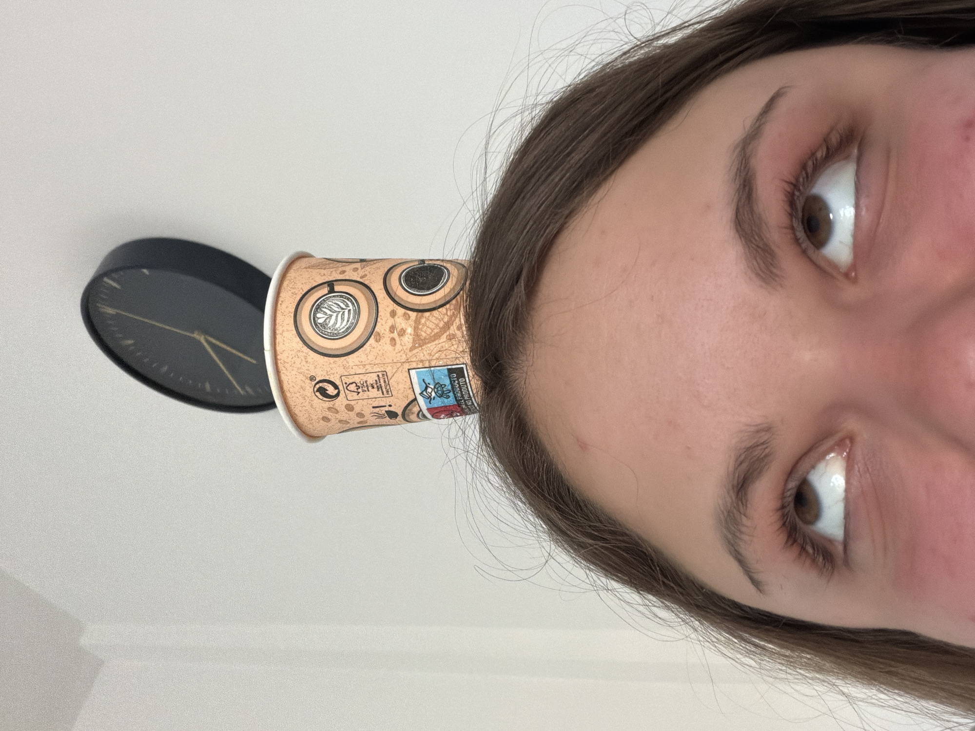 A close-up, rotated image shows a person's face with a cardboard coffee cup positioned on their forehead, seemingly balanced there. To the left of the cup is a black wall clock. The person has light brown hair and brown eyes. The background appears to be a plain, white wall. The image does not provide enough information to determine the location is Arroyo de la Miel-Benalmádena Costa, Spain.