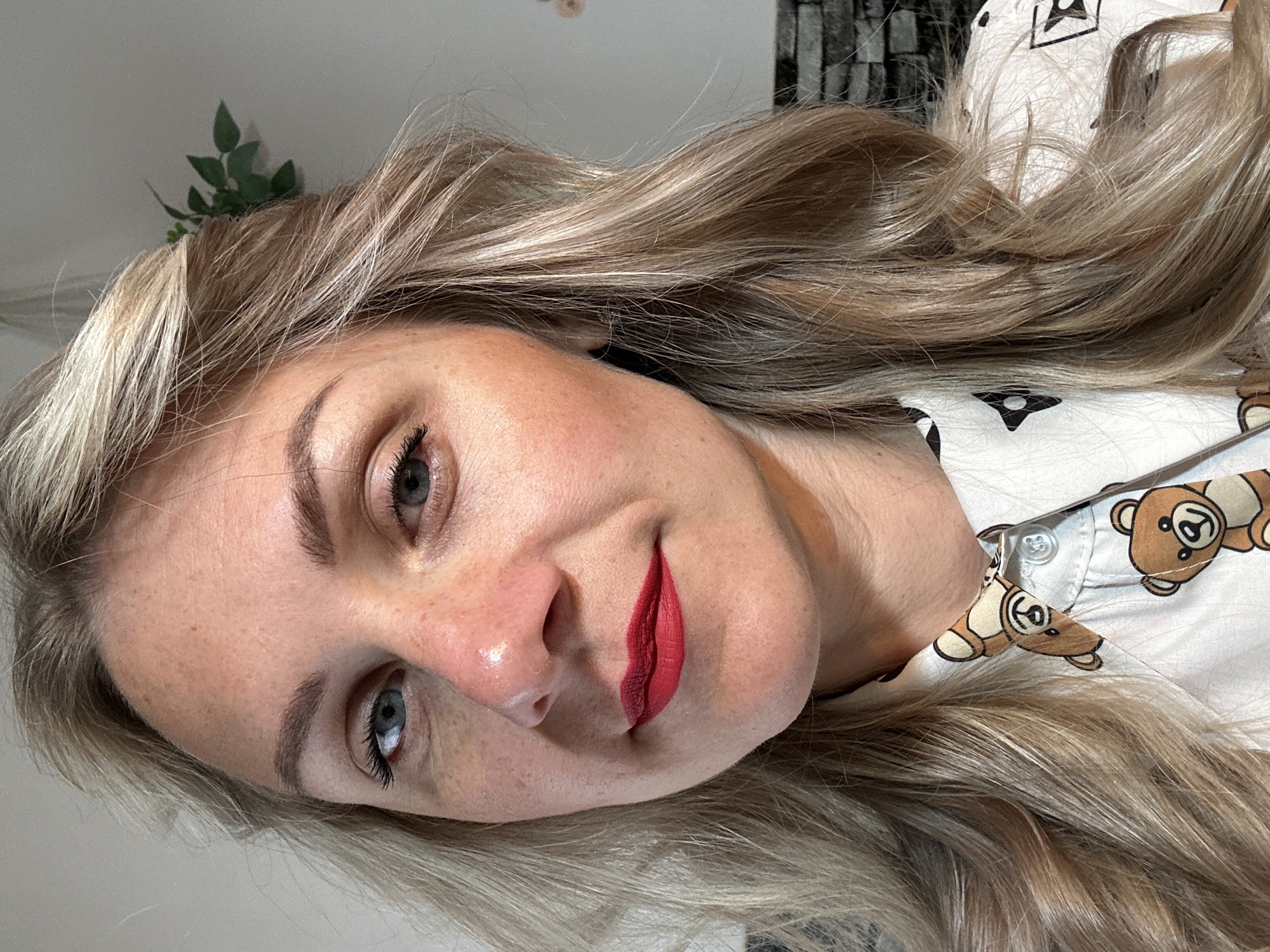 A woman with blonde hair, blue eyes, and red lipstick is depicted in a close-up shot. She is wearing a white shirt with teddy bear motifs. A small green plant is visible in the background. The location is unknown; it is not possible to ascertain whether the image was taken in Sesto Campano, Italy.