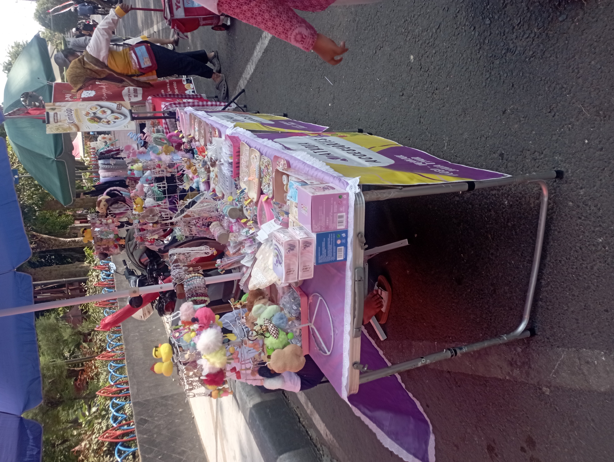 A street vendor in Nganjuk, Indonesia, displays an assortment of accessories on a folding table covered with a purple tablecloth. The stall is shaded by a blue umbrella and a green canopy. Various trinkets, including keychains, hair accessories, and small toys, are arranged on racks and in containers. A motorcycle is parked behind the stall. In the background, a woman wearing a hijab and a yellow top is seen pushing a red cart. Other people are visible in the distance, suggesting a public area. The ground is paved with asphalt.