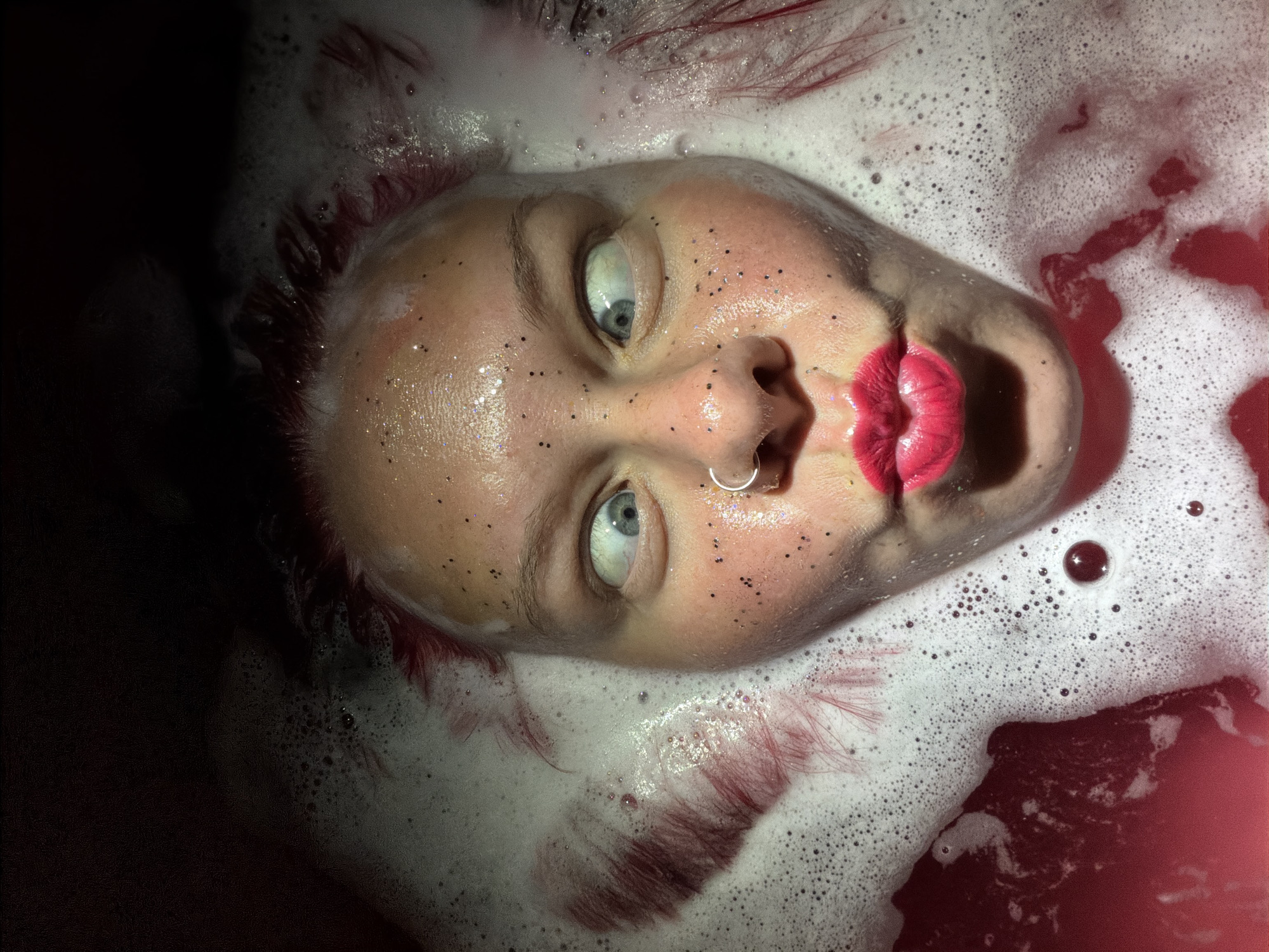 A close-up, high-angle portrait captures a person's face partially submerged in red-tinted water, surrounded by abundant white foam, in Unknown, Unknown. The individual, with fair skin, displays a puckered mouth with bright pink/red lipstick. Their blue eyes are wide open and severely crossed, looking inward. A silver hoop nose ring is visible on the left nostril. Numerous small, dark, reflective specks are scattered across the forehead, cheeks, and nose. Wet, reddish hair strands are intermingled with the foam on either side of the face. The area above the head is an indistinct, dark surface.