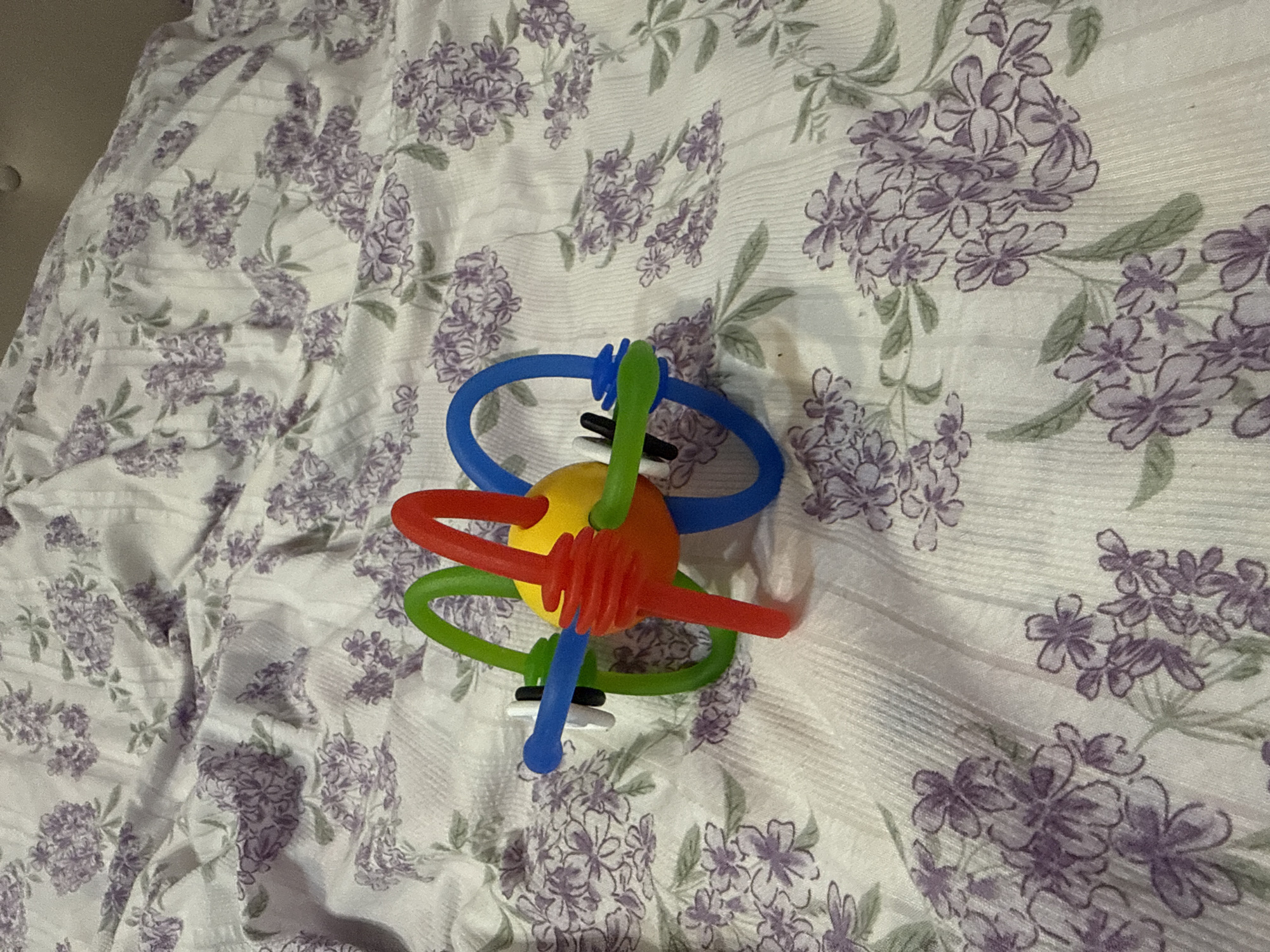 A toy rattle is placed on a white blanket with a floral pattern of purple flowers and green leaves. The rattle is a spherical, yellow center with several colorful loops extending from it. The loops are red, blue, and green, and some have ribbed textures. The toy appears to be made of flexible plastic and is positioned in the center of the frame. The lighting suggests indoor illumination. The scene is still and no activity is taking place. There is no visible text in the image. The location is identified as Pushkino, Russia.