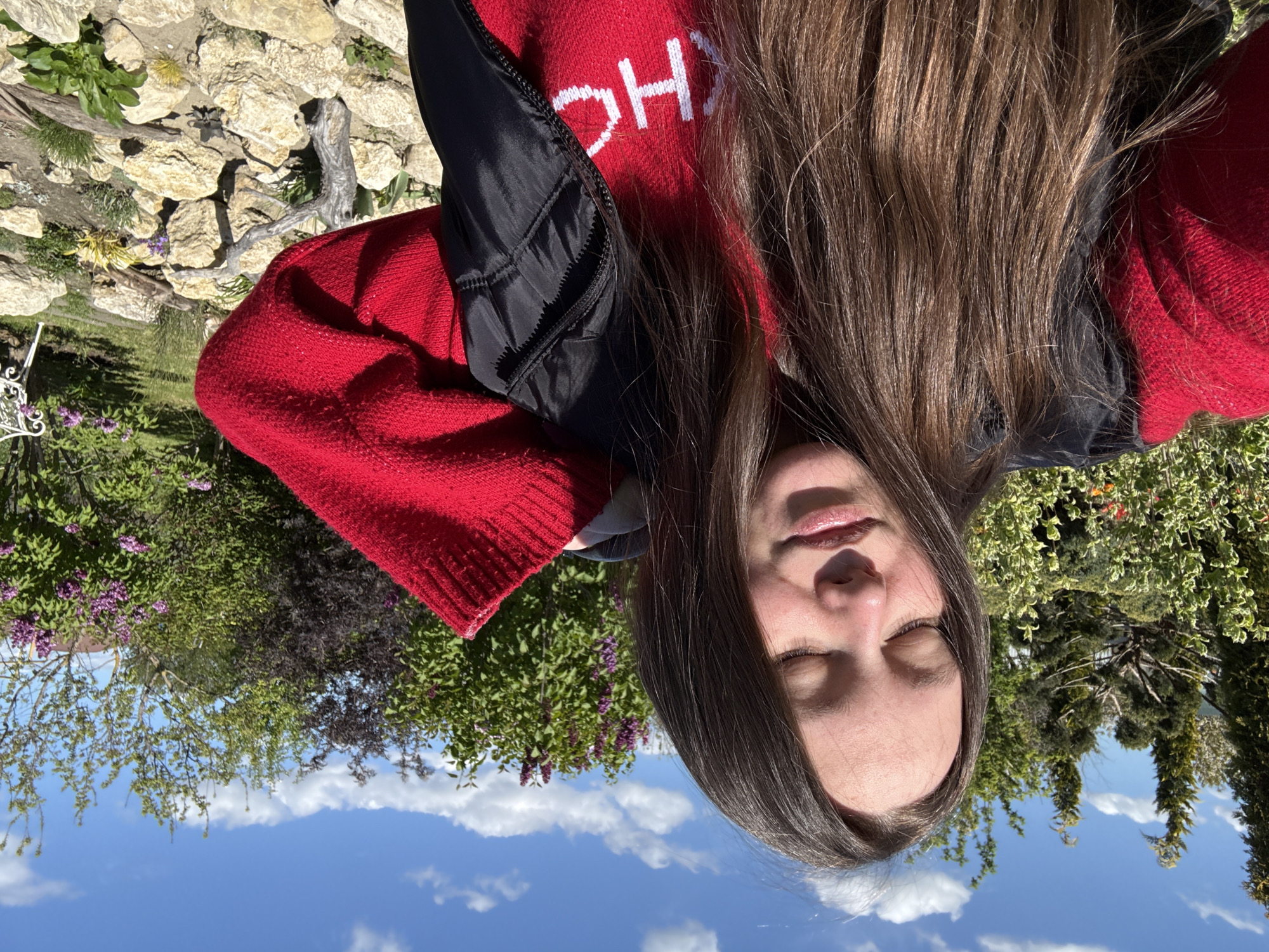 This image captures a close-up, high-angle portrait of a young person lying outdoors, looking upwards towards a bright sky, with the photo oriented 90 degrees counter-clockwise.
The subject has long, dark brown hair that cascades around their head and shoulders. Their eyes are gently closed, conveying a sense of relaxation or contentment. They are wearing a vibrant red knitted sweater, with partial white lettering, "OH", visible on the chest. A black puffer vest or jacket is worn over the sweater.
The background reveals a garden or natural outdoor setting. To one side, there are areas of rocky ground interspersed with small green plants and dry branches. Further back, the scene is filled with lush greenery, including bushes with purple flowers, possibly lilacs, and various trees. Above the foliage, a clear blue sky stretches out, dotted with scattered white cumulus clouds.
The strong illumination suggests it is daytime, likely midday or early afternoon, on a sunny and pleasant day. The visible details of a tranquil garden with blooming flowers, combined with the subject's relaxed posture, evoke a peaceful moment spent enjoying nature. The location context is Pervorechenskoye, Russia.