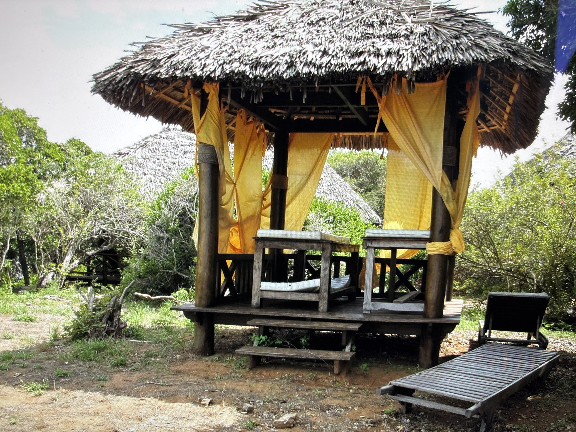 An outdoor gazebo with a thatched roof and yellow curtains is set in a natural environment with trees and foliage. The gazebo is supported by wooden posts and has a wooden deck with benches. In front of the gazebo are two wooden lounge chairs. The ground is covered with dirt and dry leaves. There are no people in the image. The scene does not appear to be in Sofia, Bulgaria.