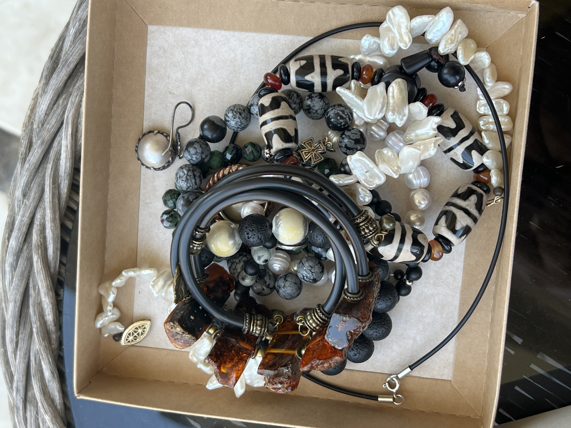 A shallow, open, light brown cardboard box is filled with a variety of necklaces and bracelets. The jewelry consists of beads made from materials such as black lava rock, faceted gray and white stones, dark green stones, creamy white irregular pearls, and chunky, translucent amber-colored pieces. There are also several larger, cylindrical beads with black and white geometric patterns, and a small, ornate cross-shaped charm. The jewelry is artfully arranged, with some pieces layered on top of others, and the box is positioned on a dark, reflective surface, possibly a table, with a textured, woven material visible in the background. The lighting is natural and appears to be coming from above, casting subtle shadows. There are no people or animals present in the image.