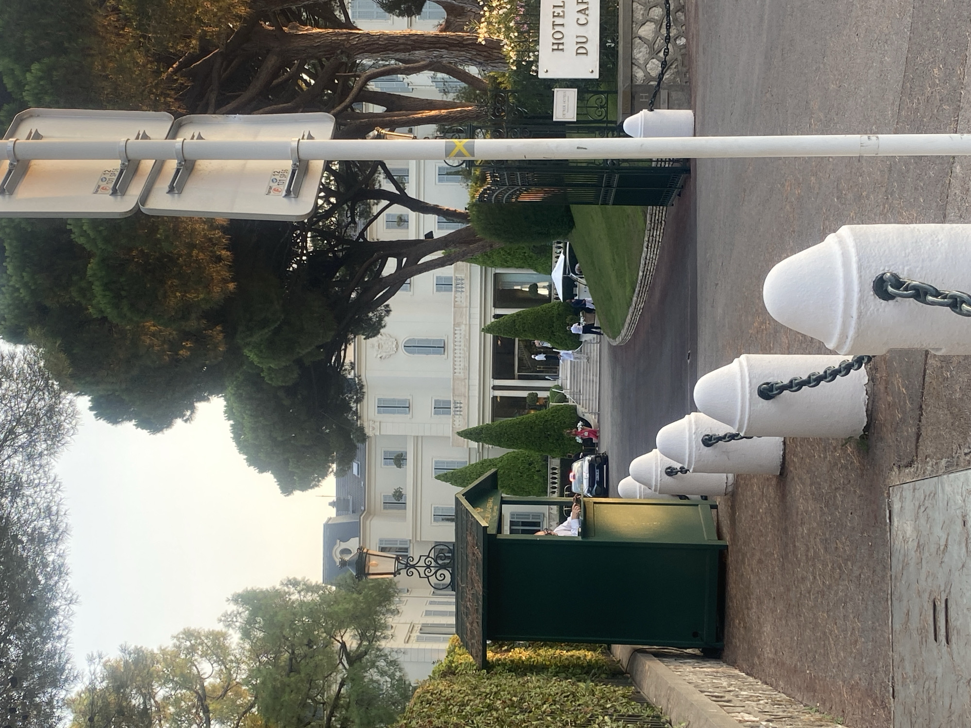 The image shows the entrance to the Hôtel du Cap-Eden-Roc in Antibes, France. The grand white hotel building is visible in the background, surrounded by mature trees. In the foreground, there is a green security booth with a person inside. White bollards connected by chains line the driveway. A signpost with traffic signs is also present. The scene appears to be set in the late afternoon or early evening, with soft sunlight illuminating the area.
