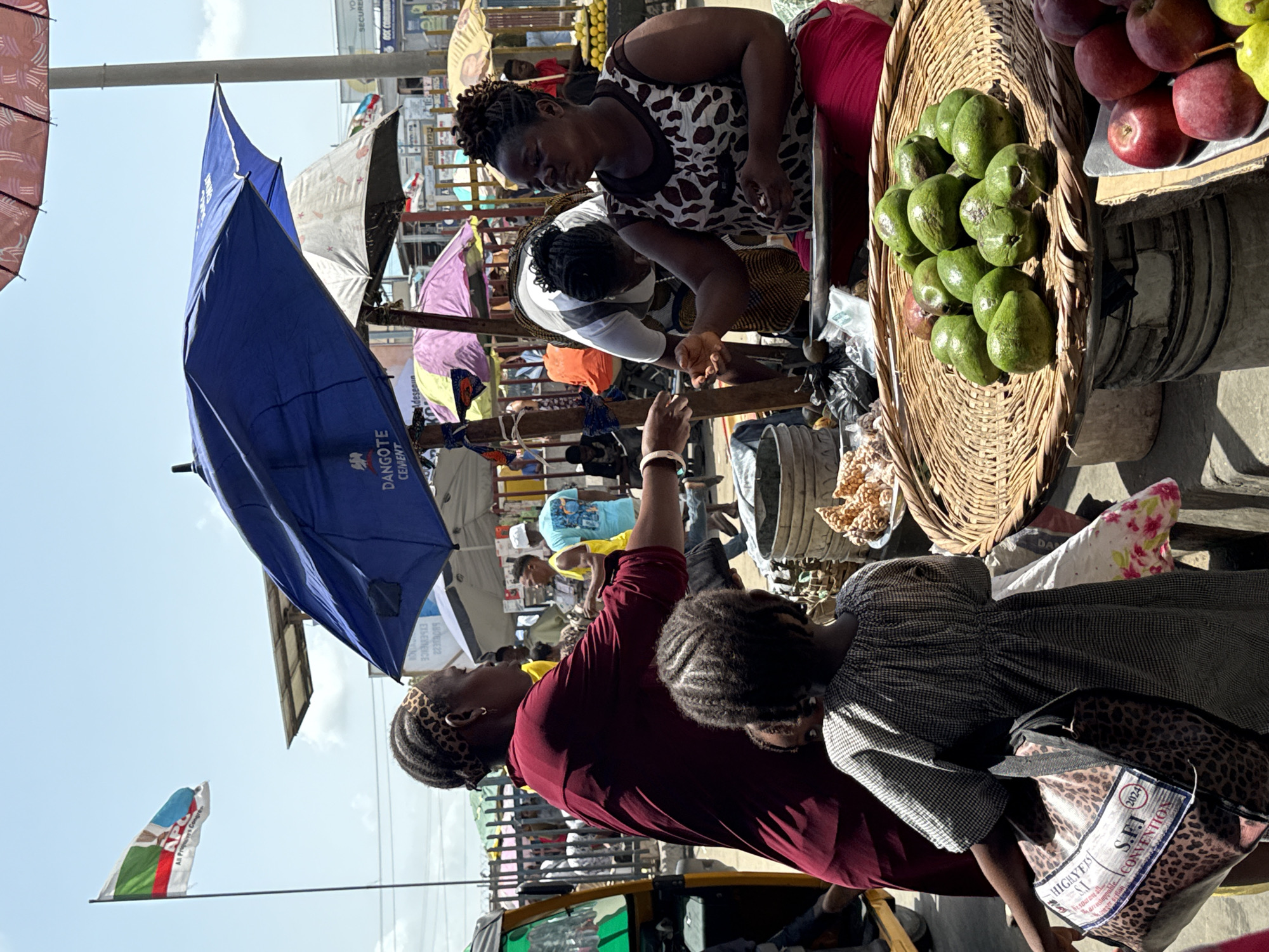 This outdoor scene captures a bustling marketplace in Lekki, Nigeria. A woman in a maroon top and a child are in the foreground, seemingly engaging with a vendor. Nearby, another woman in a spotted dress interacts with a customer, possibly selling goods. Baskets of avocados and apples are visible on a table. Several market umbrellas provide shade, with one blue umbrella displaying "DANGOTE CEMENT" branding. In the background, more people are visible amidst the market activity. A flag with "APC All Progre Congres" is also seen, alongside a bag reading "S.F.J. (2024) CONVENTION".