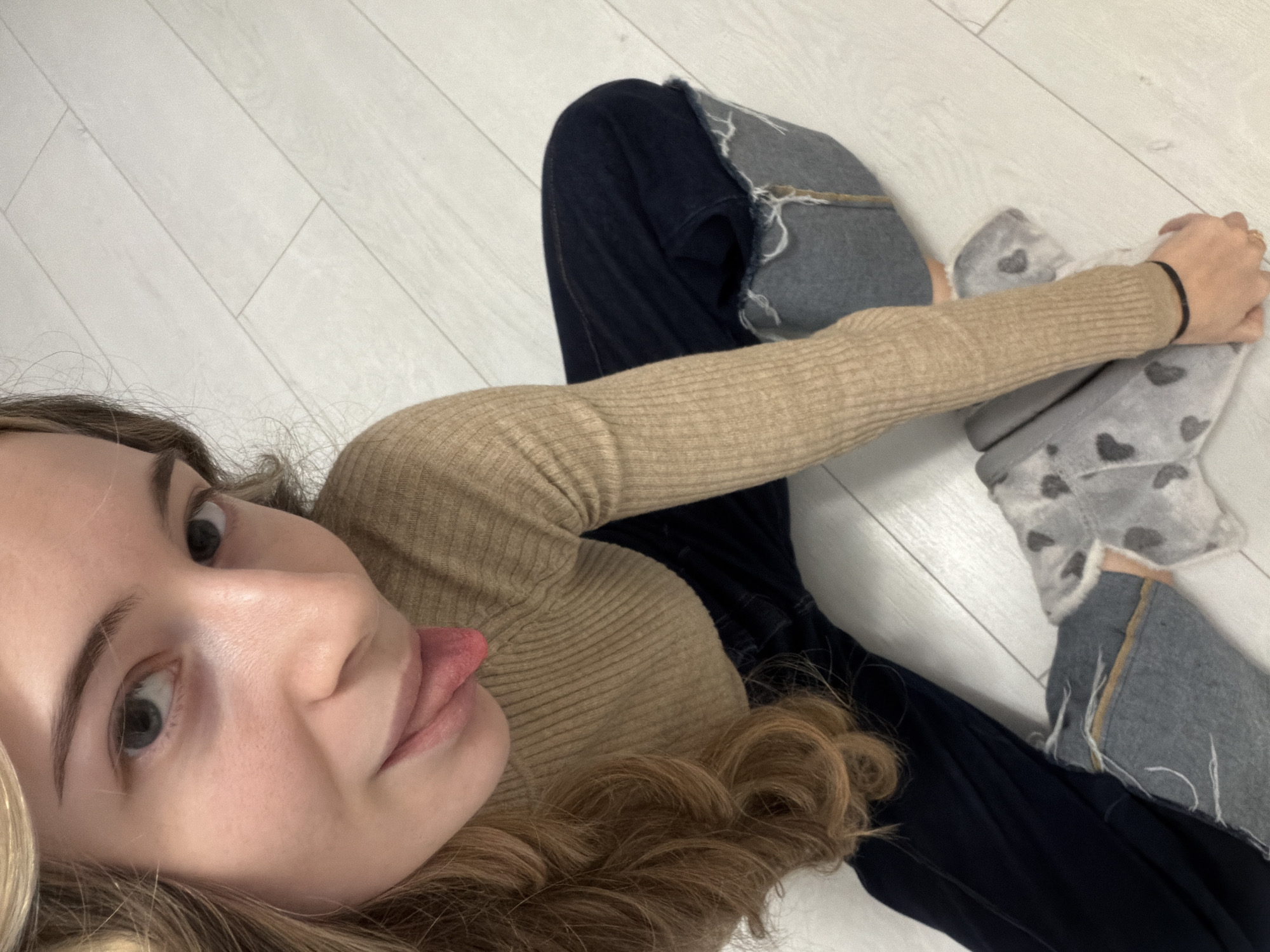 A young woman is seen from an elevated, angled perspective lying on a light-colored, planked floor. She is wearing a tan-colored, long-sleeved, ribbed shirt and dark blue jeans with ripped denim patches on the knees. Her feet are covered in gray slippers adorned with black heart patterns. Her left arm is extended toward her feet, and she is holding the slippers with her left hand. A black hair tie is visible on her left wrist. Her face is angled towards the camera, and she is sticking her tongue out. Her hair is blonde and wavy. The background is limited to the white floor. The location is not identifiable as Plovdiv, Bulgaria, based on the provided visual information.