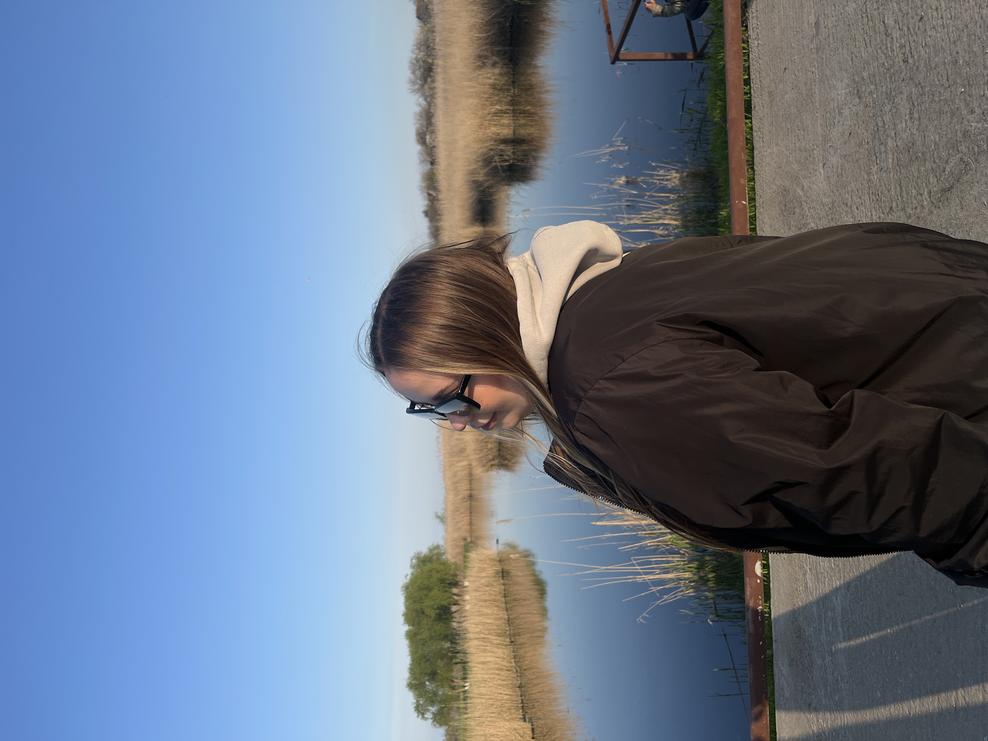 A sideways portrait captures a young person, likely female, positioned on a paved embankment or pier next to a body of water. The subject has light brown or blonde hair and wears dark sunglasses, a light-colored hoodie, and a dark, possibly black, jacket. They are looking downwards, with their face partially obscured, suggesting quiet observation of the scene below or in front of them.
To the right of the person, a grey concrete surface and a dark, possibly metal, railing or structure are visible. The background features a calm body of water, likely a river or canal, which reflects the clear blue sky and the opposite bank. The far bank is characterized by vast stretches of dry, tall grasses or reeds, with a cluster of green trees visible further in the distance. The sky above is a bright, clear blue, indicating good weather and daytime conditions with soft, natural lighting. No specific activity beyond observation is evident, and no visible text is present in the image. The location context is Novopokrovskaya, Russia.