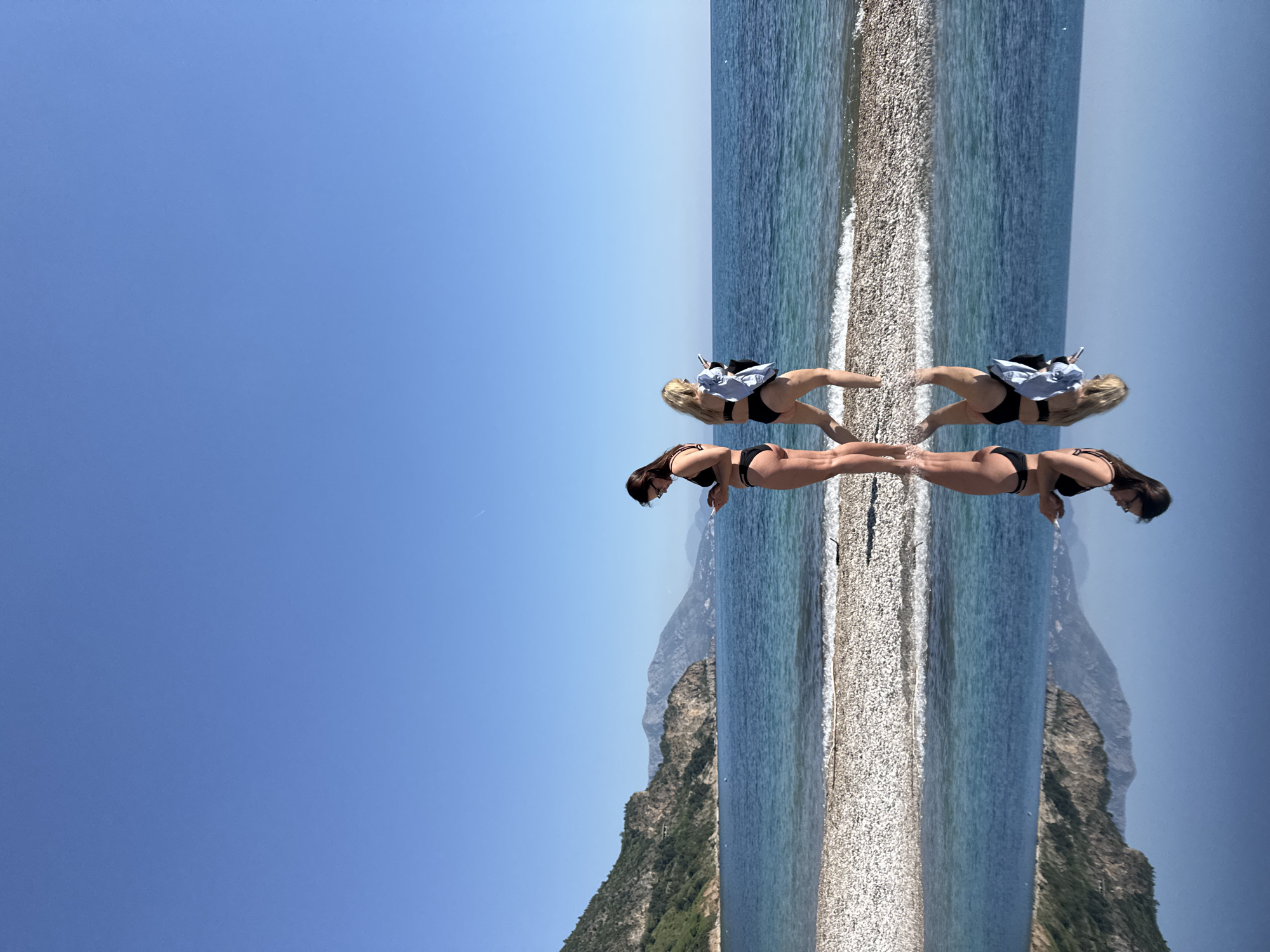 The image is a vertically mirrored view of a coastal scene, likely located in Prijevor, Montenegro, given the context.
The primary subject is a pair of women on a pebble beach adjacent to the sea. They are dressed in swimwear. The image is mirrored, creating a symmetrical effect around the central line of the beach.
The background consists of the sea and a mountain terrain with greenery. The sky is visible in the upper portions of the mirrored image, showing a clear, blue expanse.
The action depicted involves the women seemingly interacting on the beach. Their interaction is mirrored as well, reinforcing the symmetry of the image.