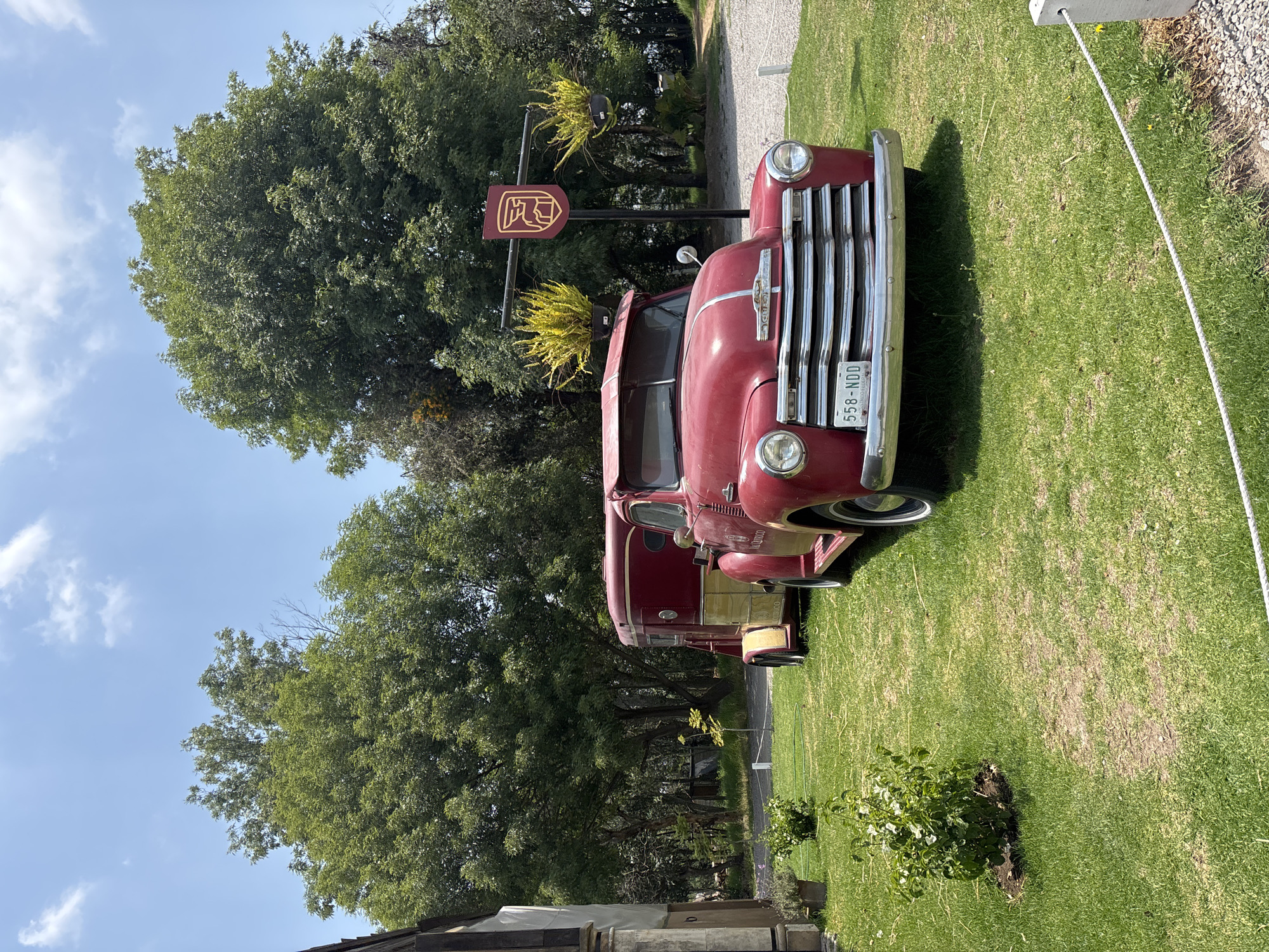 A red, vintage Chevrolet truck is parked on a grassy lawn in Domingo Arenas, Mexico. The truck faces the viewer with its front grille prominently displayed. A sign with a coat of arms, adorned with hanging plants, stands nearby. Trees provide a backdrop to the scene, with a blue sky partially visible. A concrete post with a rope extending to the right suggests a boundary or pathway. The truck appears to be in a display setting. A partial view of a building is visible in the foreground.