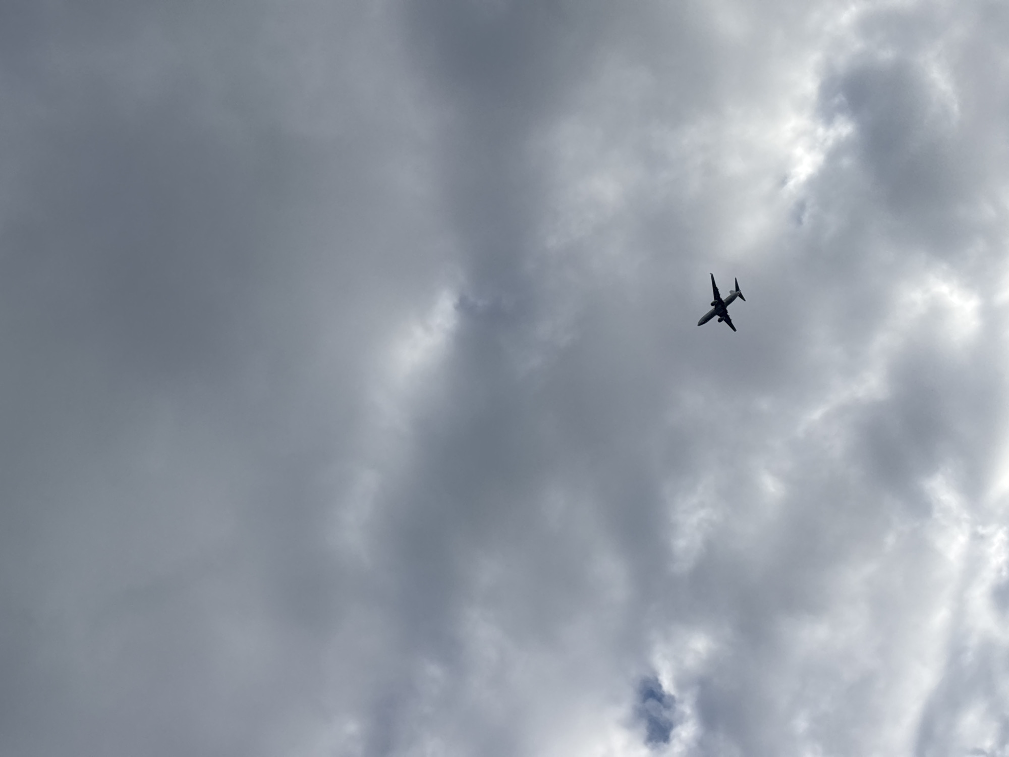 The image shows an airplane flying in a cloudy sky. The plane is silhouetted against the gray clouds and is angled upwards. The environment consists primarily of varying shades of gray clouds, with some brighter areas suggesting sunlight behind the clouds. The location within Varna, Bulgaria, cannot be determined from the image content.