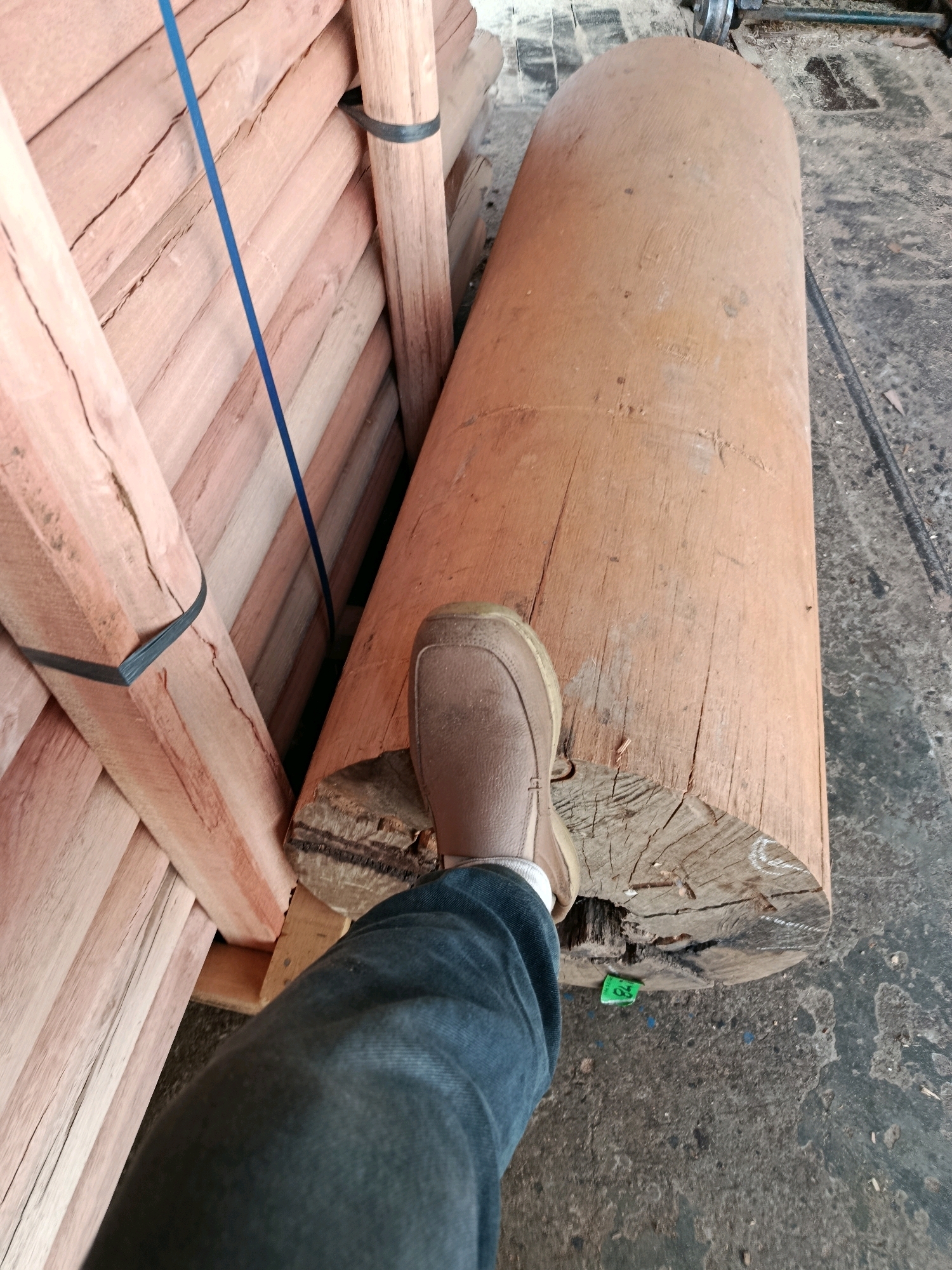 In the city of Kembangarum, Indonesia, a person's lower body is visible in the foreground of an outdoor scene. They are wearing dark blue jeans and a light brown shoe. The person's right foot is positioned on a large, rough-hewn log that has a cracked cross-section. Behind the log, to the left, is a stack of reddish-brown wooden planks, bound together with a dark strap and a blue string. The ground beneath the log and planks appears to be concrete or asphalt, with some debris scattered on it. The overall impression is of a storage or work area, possibly a lumber yard or construction site.