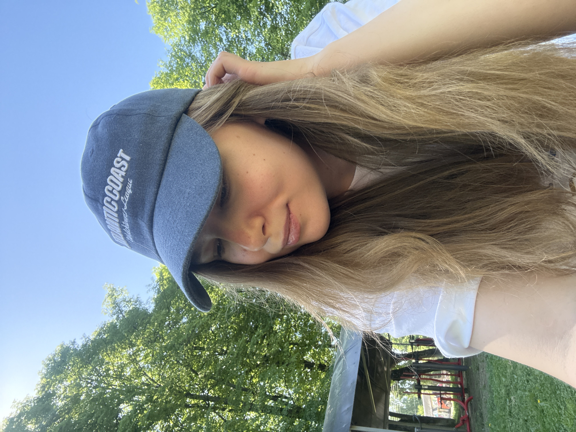 A young woman wearing a blue cap with white lettering is pictured from a low angle under a clear blue sky. She is wearing a white t-shirt and has long brown hair. Her hand is touching the top of her head. The background includes green trees and grass, suggesting she is outdoors, possibly in a park. Some metal structures with red accents are visible in the background as well. The location is Красный Маяк, Russia.