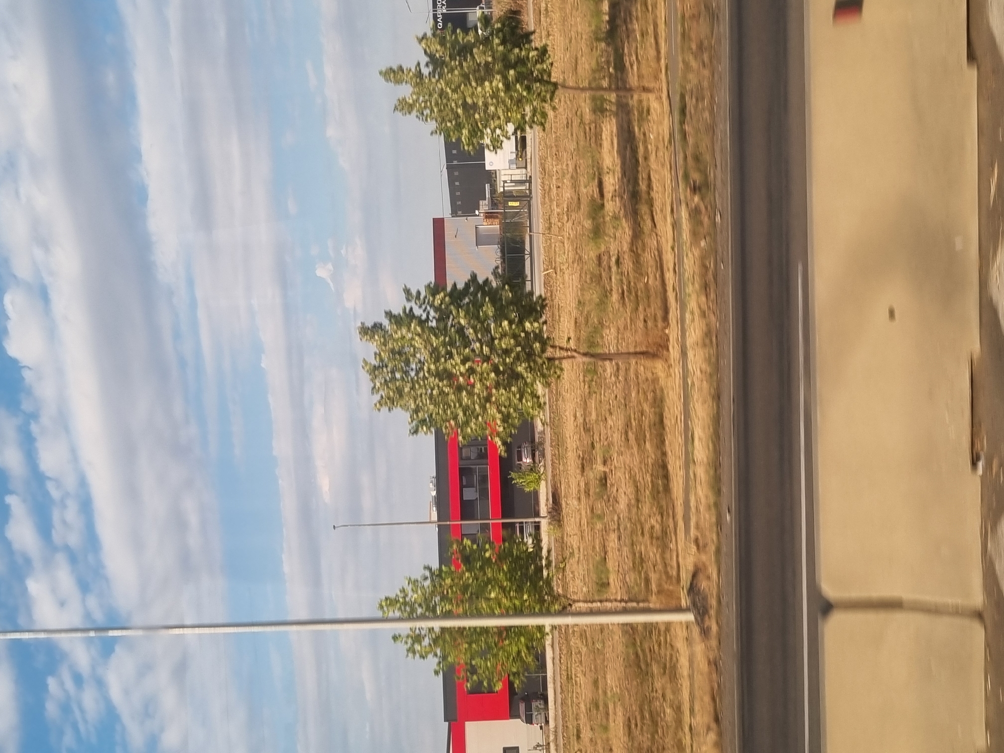 The image shows a street view in Sântion, Romania, on a sunny day with scattered clouds. In the foreground, a concrete barrier is visible. Beyond the barrier is a road with a dividing line, and then a dry, grassy field. Several trees with green foliage are planted in the field. In the middle ground, a modern building complex is visible. One section of the building is red and white, with dark windows. Another section of the building is dark with multiple windows. A tall, thin pole stands to the left of the building. In the background, the sky is blue with white clouds. Several vehicles are parked in front of the building complex, including what appear to be vans and cars. The scene appears to be an industrial or commercial area.