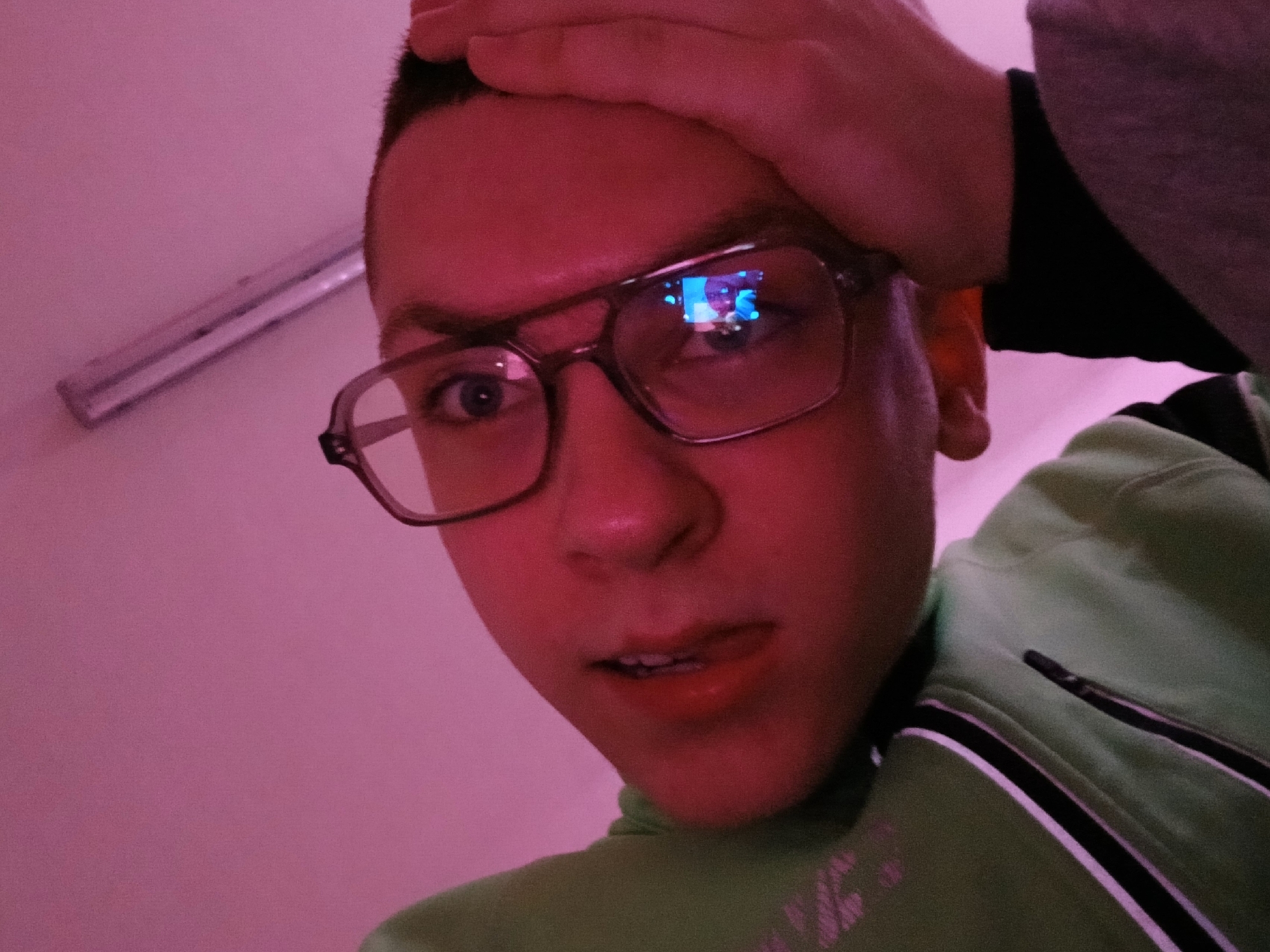 A close-up, low-angle shot depicts a person with fair skin, short, dark hair, and wearing glasses. The person's hand is touching their forehead. A reflection is visible in the person's glasses. The person is wearing a light green jacket with a white and black stripe. The wall is pink. A light fixture is visible in the upper left corner. The location and city is not identifiable from this image.