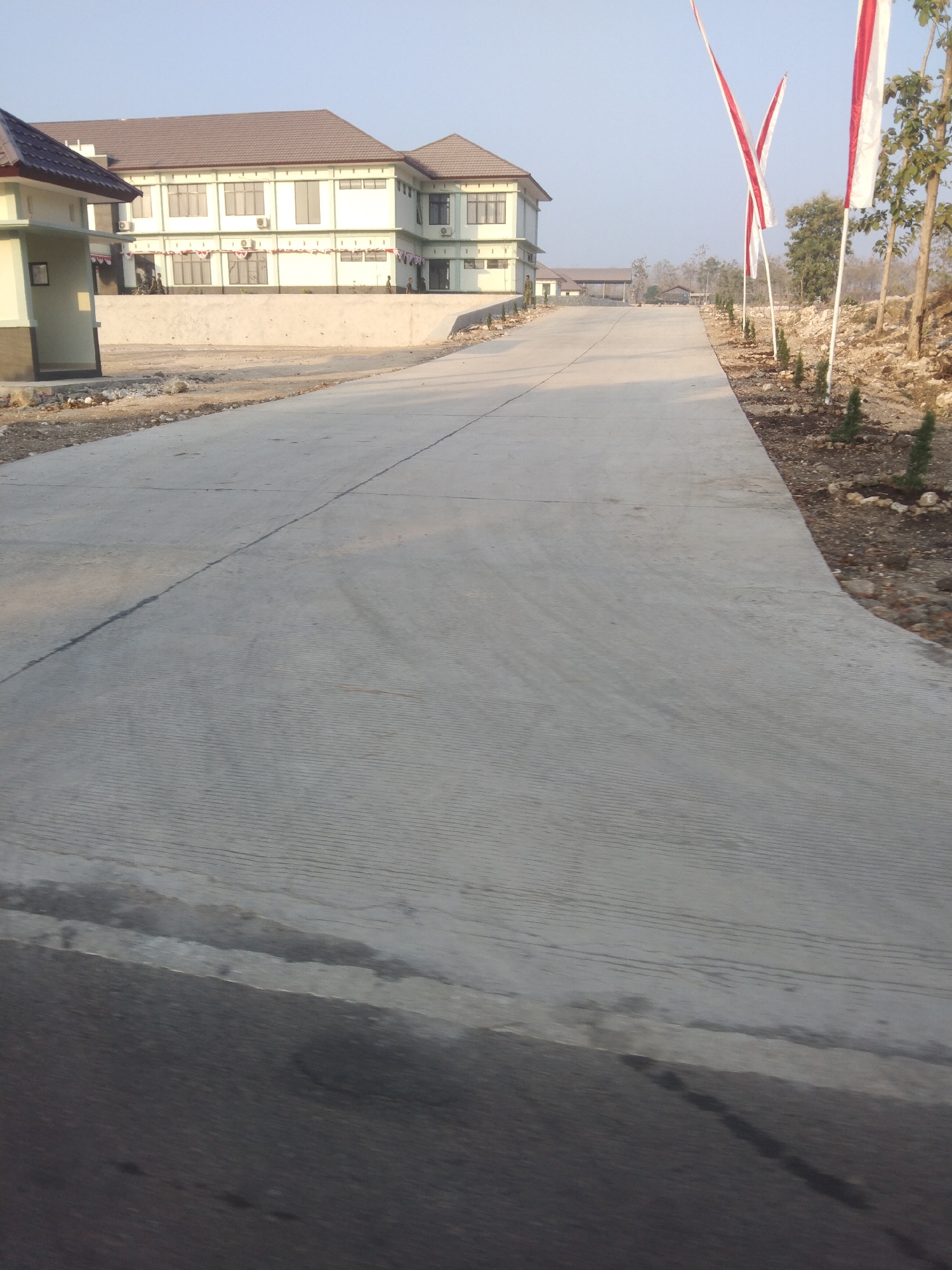 The image depicts a concrete road leading towards a light-colored building with a brown roof, situated in the city of Growok, Indonesia. The building appears to be a school or administrative complex, with multiple windows and air conditioning units visible. Along the right side of the road, three flags with red and white stripes are planted, fluttering in the breeze. The area surrounding the road is sparsely vegetated, with some small trees and rocky terrain. The sky is clear and the overall lighting suggests it is daytime, possibly morning or late afternoon, given the soft shadows. A small, light-colored structure with a brown roof is visible on the left side of the road, closer to the foreground. There are a few indistinct human figures near the building. The road itself has visible expansion joints and a textured surface.