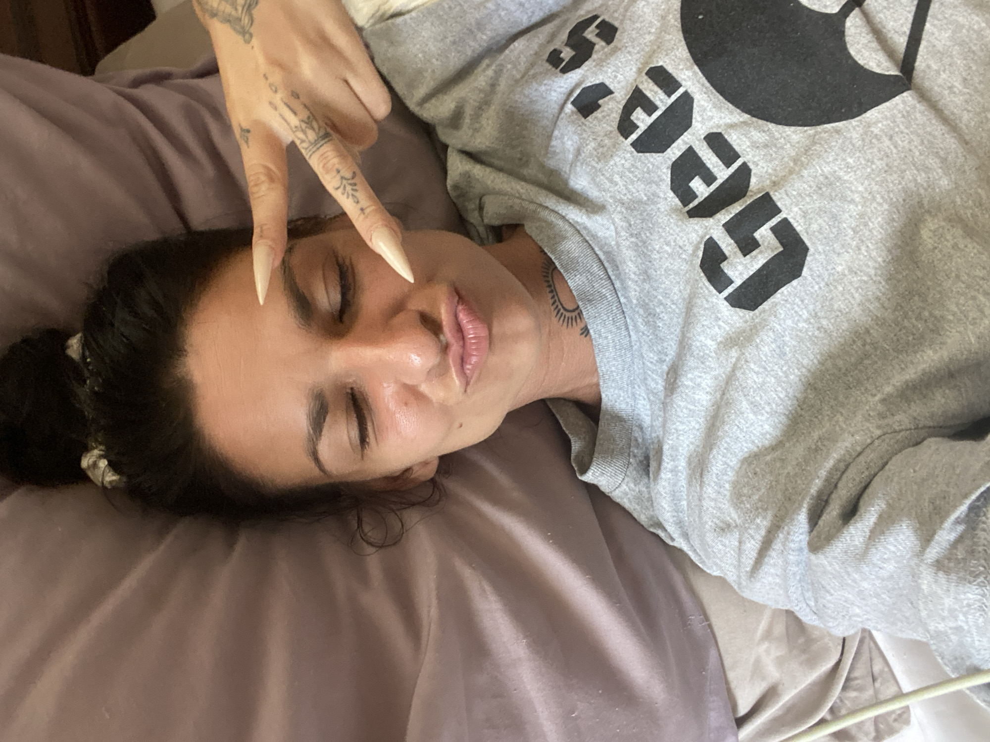 A woman is lying in bed with her eyes closed and her lips puckered. She is wearing a grey sweatshirt with the words "Gee's" printed on it. Her hand is raised to her face, and she is making a peace sign with two fingers. There are tattoos on her fingers and neck. The bed is covered in a light purple sheet. The scene appears to be indoors. This image does not contain any identifiable landmarks or contextual information to suggest a location in Mũi Né, Vietnam.