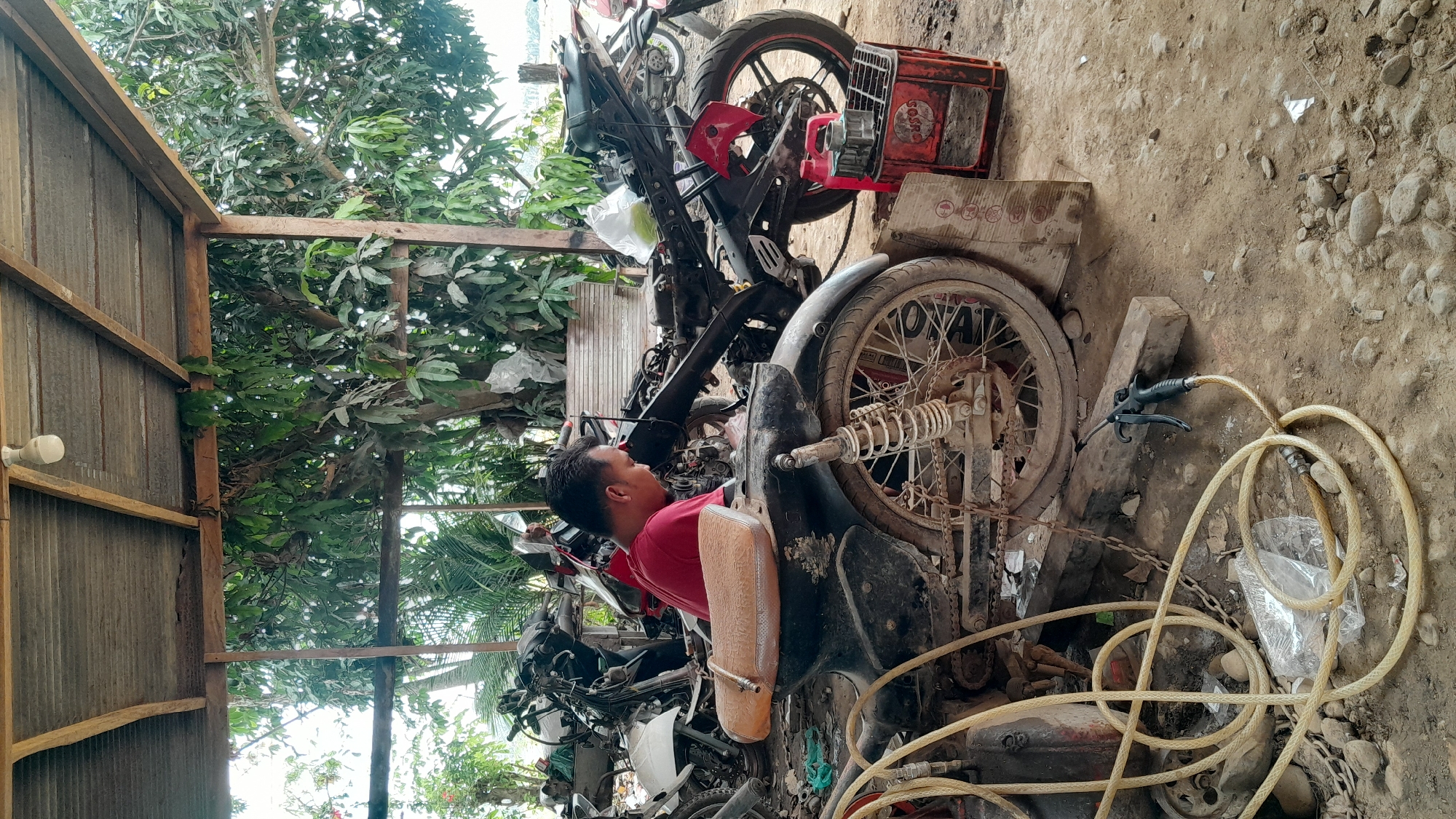The image shows a man in a red shirt working on a motorcycle in Lhokjok, Indonesia. The motorcycle is positioned in a cluttered outdoor area with various motorcycle parts scattered around. The area is covered by a corrugated metal roof supported by wooden frames. A tree with lush green foliage is visible in the background. The ground appears to be a mix of dirt and rocks. In the foreground, there is a yellow air hose and some plastic bags.