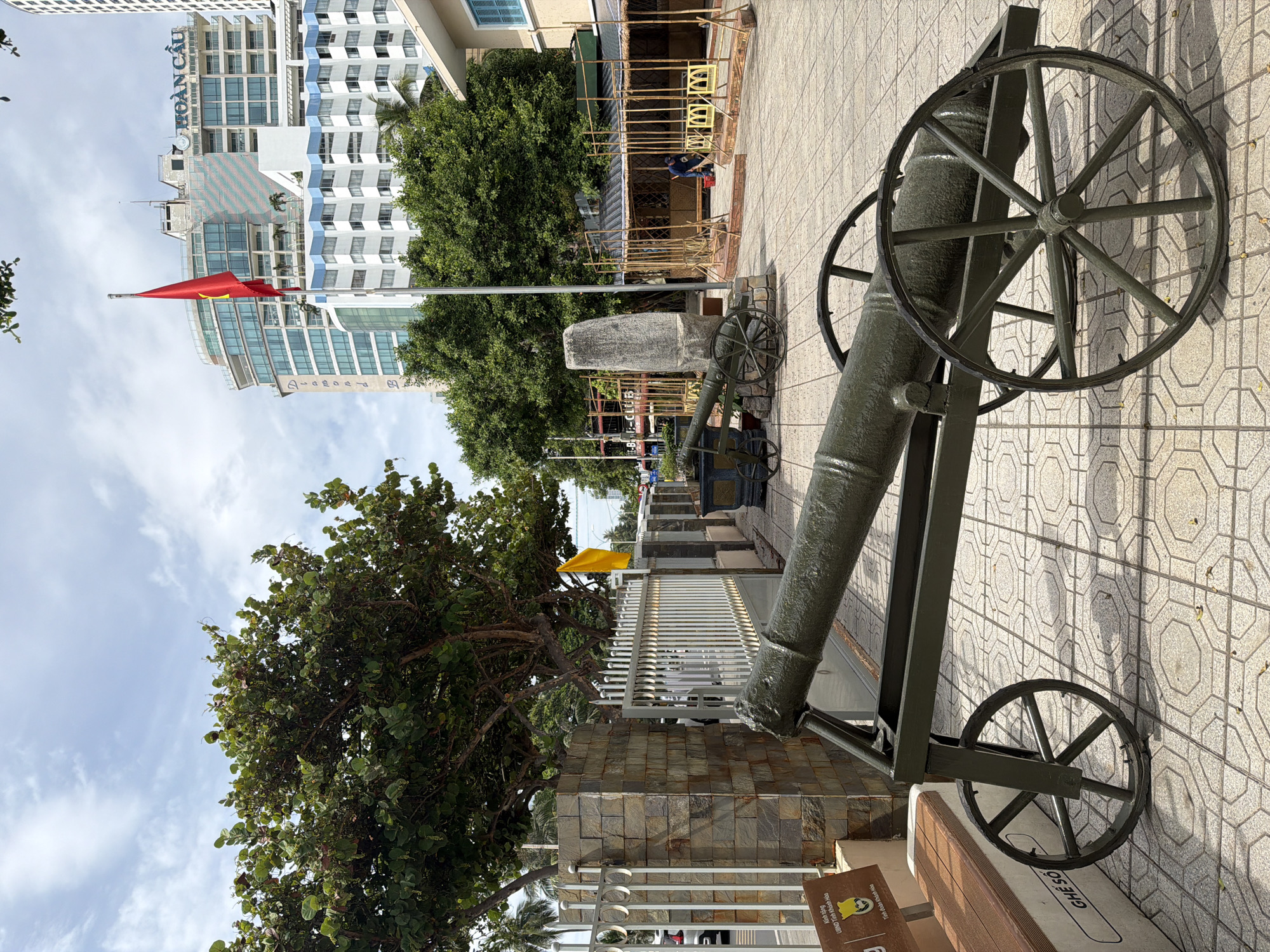 This image depicts a historical cannon on display on a tiled sidewalk in Nha Trang, Vietnam, with a modern cityscape in the background. The cannon, painted dark green, is mounted on a wheeled carriage. A flag of Vietnam is prominently displayed on a flagpole to the left of the buildings. The buildings are modern, with glass facades and balconies, and one building is labeled "HOÀN CẦU" which translates to "Globe" or "Global." There is lush greenery in the foreground, including a large tree and some smaller bushes. A stone monument stands behind the cannon. In the middle ground, there are temporary structures made of bamboo. A person wearing a blue shirt and dark pants is visible in the background, appearing to be cleaning or working near the bamboo structures. A bench is also visible in the lower left of the image with text that appears to be Vietnamese. The sky is partly cloudy. The overall scene suggests a historical display or museum area integrated into the urban environment of Nha Trang.