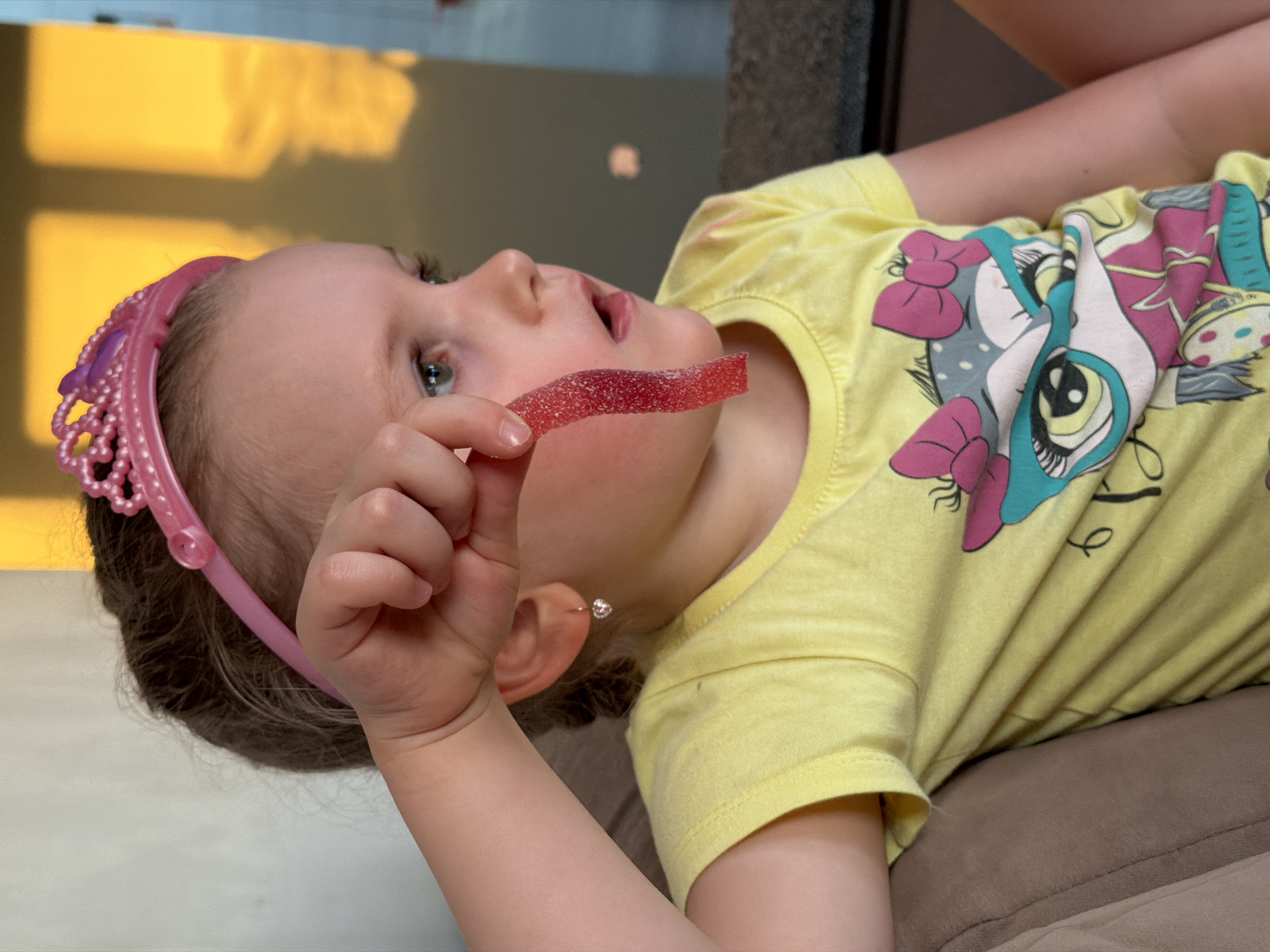 A young girl wearing a yellow My Little Pony t-shirt and a pink tiara holds a red, sugary candy strip. She is lying down, possibly on a pillow or soft surface. The location and context of the image within the city of Chaikovsky, Russia are not provided.