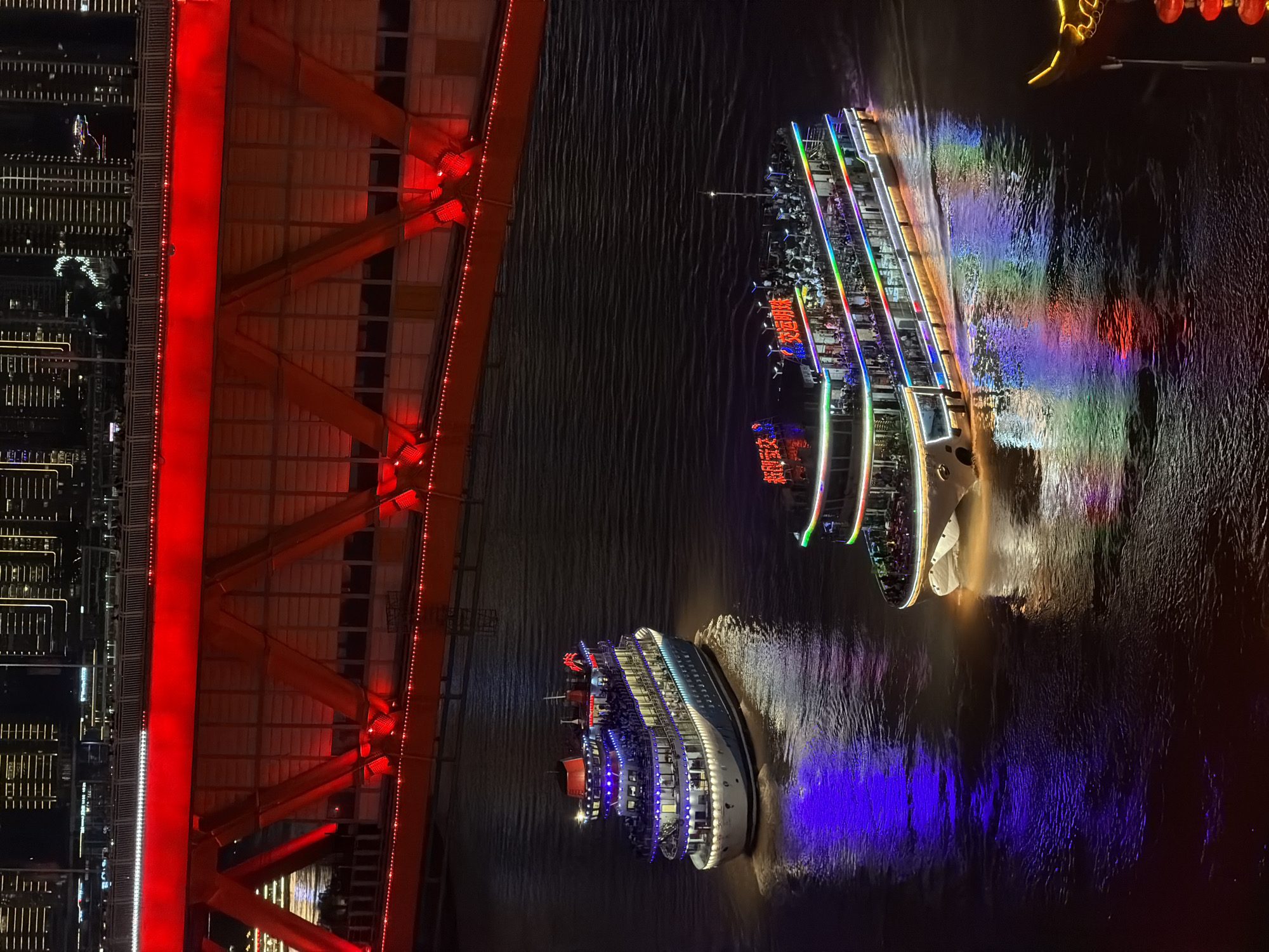 A high-angle, long shot captures a nighttime cityscape in Yuzhong District, China, featuring two illuminated tour boats navigating a river beneath a brightly lit red bridge. The bridge spans the river, its red lights creating a dramatic contrast against the dark water and the silhouetted buildings in the background. The two boats, filled with visible passengers, are adorned with vibrant LED lights in blue, green, red, and yellow, casting colorful reflections on the rippling water. The boat closer to the foreground is larger and more elaborately decorated. The city skyline, composed of numerous tall buildings with illuminated windows, forms the backdrop. The overall scene depicts a lively urban night, with the illuminated boats and bridge serving as focal points.
