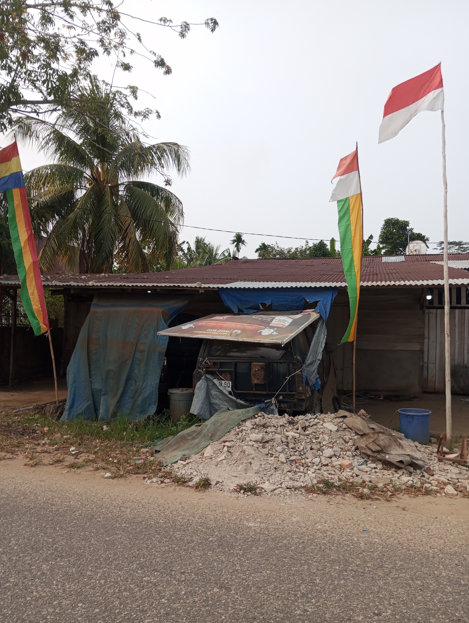 In Pematang Reba, Indonesia, a roadside scene unfolds under an overcast sky. The foreground is dominated by an asphalt road, with a textured surface and a light brown dirt shoulder. Beyond the shoulder lies a pile of rubble and debris, a mix of concrete fragments, dirt, and scattered vegetation.
To the left, a flag with vertical stripes of red, yellow, blue, and green flutters on a wooden pole. Behind it, a palm tree with abundant fronds stands tall. Adjacent to the debris, a dilapidated structure with a corrugated tin roof is partially covered by a large blue tarp. A makeshift awning, also covered by a tarp, extends from the structure, sheltering what appears to be a truck or vehicle with a license plate reading "B 90".
Further to the right, two more flags are planted on wooden poles. One is the Indonesian flag, red and white, flying prominently. To its left and slightly lower, another flag displays vertical stripes of red, white, green, and yellow. The structure itself appears to be made of wood and is partially obscured by tarps. A blue bucket rests near the right edge of the frame, close to some wooden supports. Vegetation, including trees and shrubs, is visible in the background, suggesting a semi-rural or suburban setting. The overall impression is one of a modest, possibly under-construction or neglected, roadside establishment.