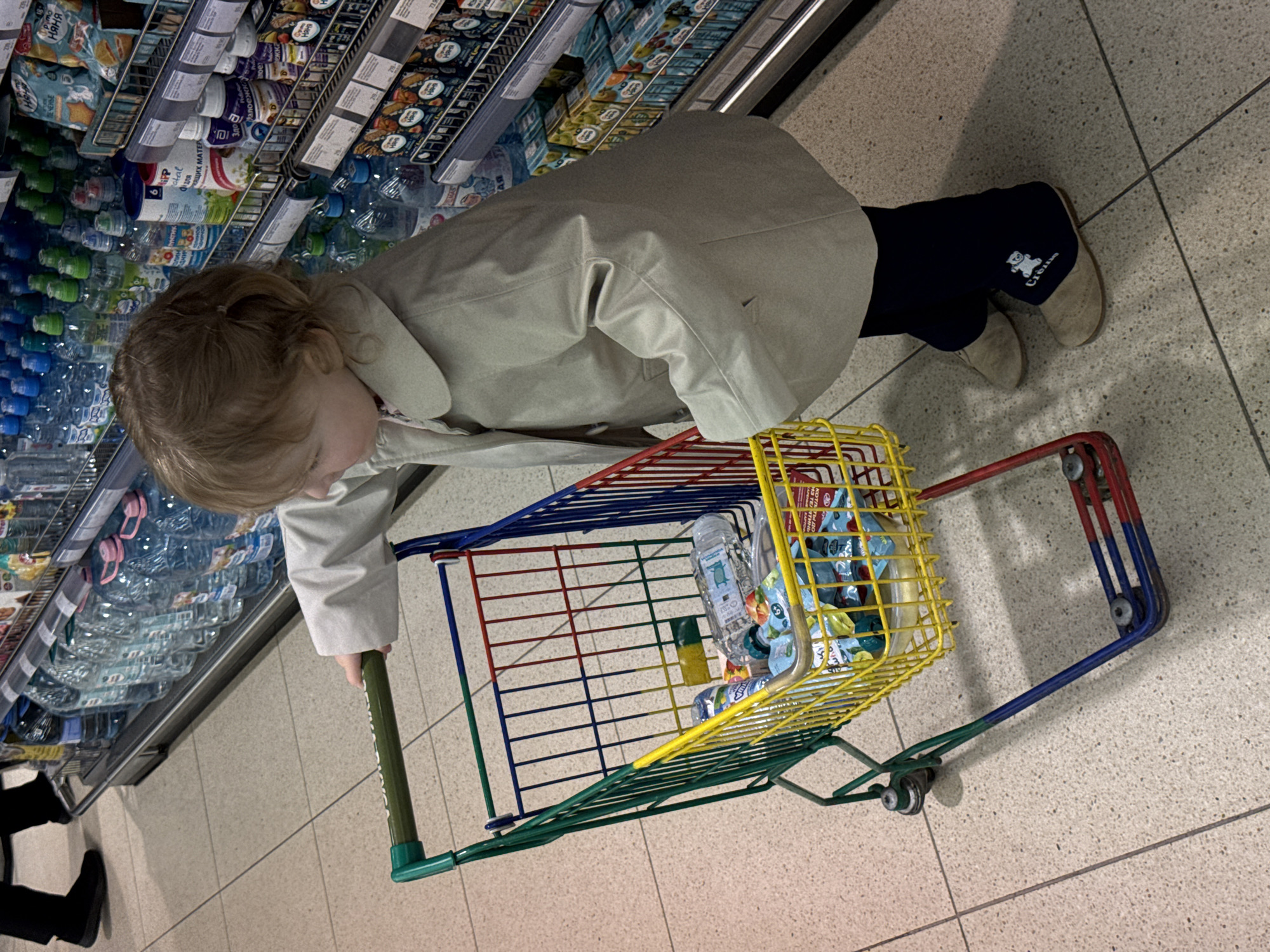 An overhead-angled photograph captures a young child in a supermarket aisle in Moscow, Russia, pushing a brightly colored mini shopping cart. The child, with light brown hair tied back, wears a light beige jacket, dark blue pants with a "Crème" teddy bear logo, and light-colored shoes. She is bending forward, looking intently at products displayed on a refrigerated shelf to her left.
The shelves are well-stocked with various beverages and packaged goods. The bottom shelves primarily hold numerous plastic bottles of water, some with green and some with blue caps, and larger multi-packs. Above these, there are shelves with smaller bottles and pouches, likely baby food or drinks, featuring Cyrillic text. Visible text on product labels includes "Няня" (Nanny), "ПЕЧЕНЬЕ" (Cookies), and "для КОРМЯЩИХ МАТЕР" (for NURSING MOTHERS), along with brand names like "Малоежка" (Maloeshka). Price tags or product codes like "918," "719," and "6" are also visible.
The child's small shopping cart is a vibrant mix of green, red, blue, and yellow bars. Inside, there are a few items, including small packaged snacks or drinks similar to those on the shelves, and at least one plastic bottle of water. The floor is light-colored tiled, segmented by dark grey grout lines. In the bottom left corner, the legs and black shoes of another person are partially visible, suggesting an adult accompanying the child on their shopping trip. The overall scene depicts a typical shopping experience from a child's perspective.