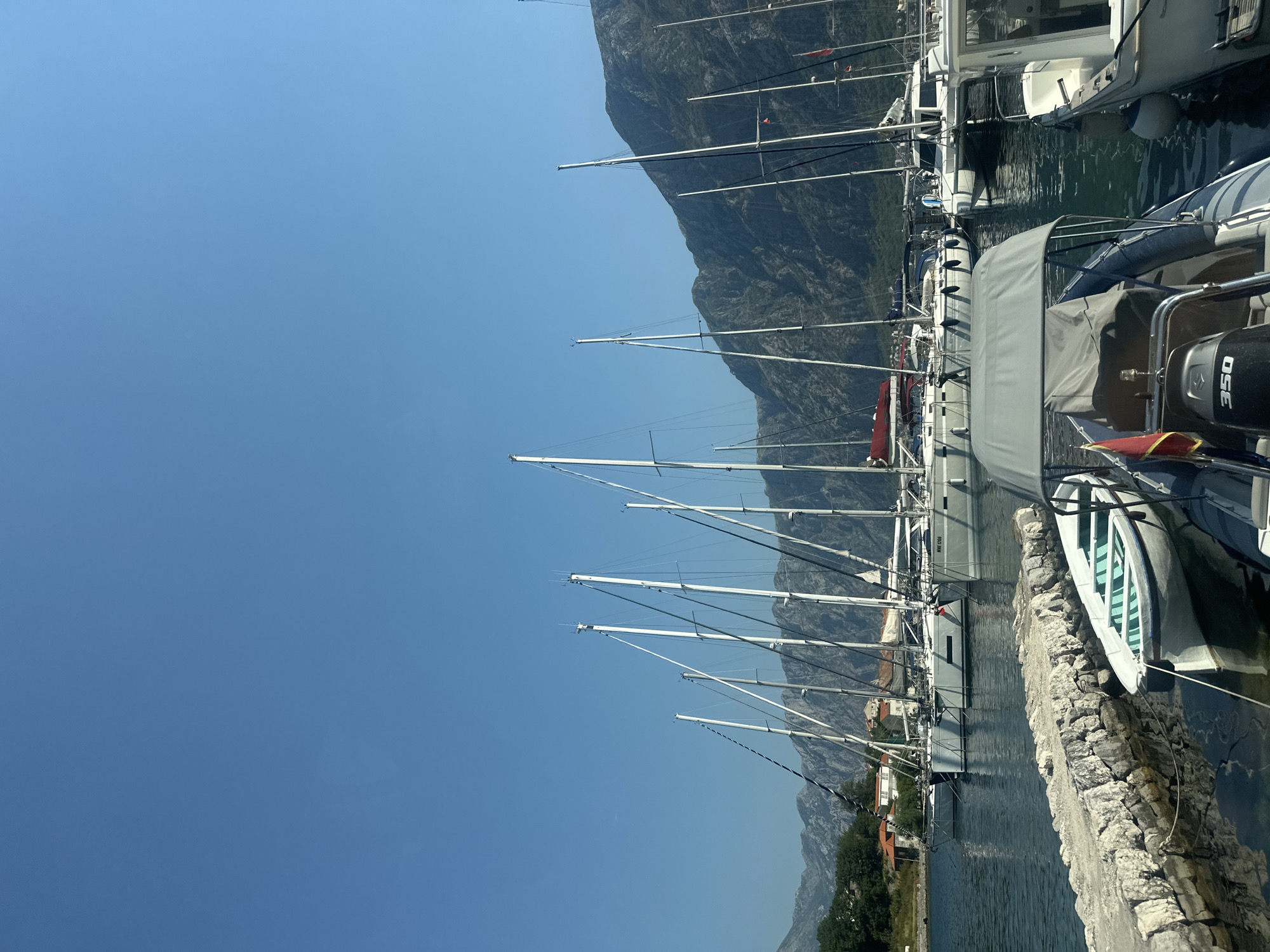 In Prčanj, Montenegro, a vibrant marina is captured under a clear blue sky. Numerous sailboats are docked, their tall masts reaching towards the heavens, interspersed with some smaller motorboats. In the foreground, a stone jetty is visible, with a small white boat tied to it, its interior painted in a refreshing shade of mint green. The water in the marina is a deep, inviting blue, reflecting the surrounding scenery. Behind the marina, a picturesque town with red-tiled roofs nestles against the backdrop of a majestic, rugged mountain range. The overall scene is one of tranquility and natural beauty, showcasing the charm of this coastal town.
