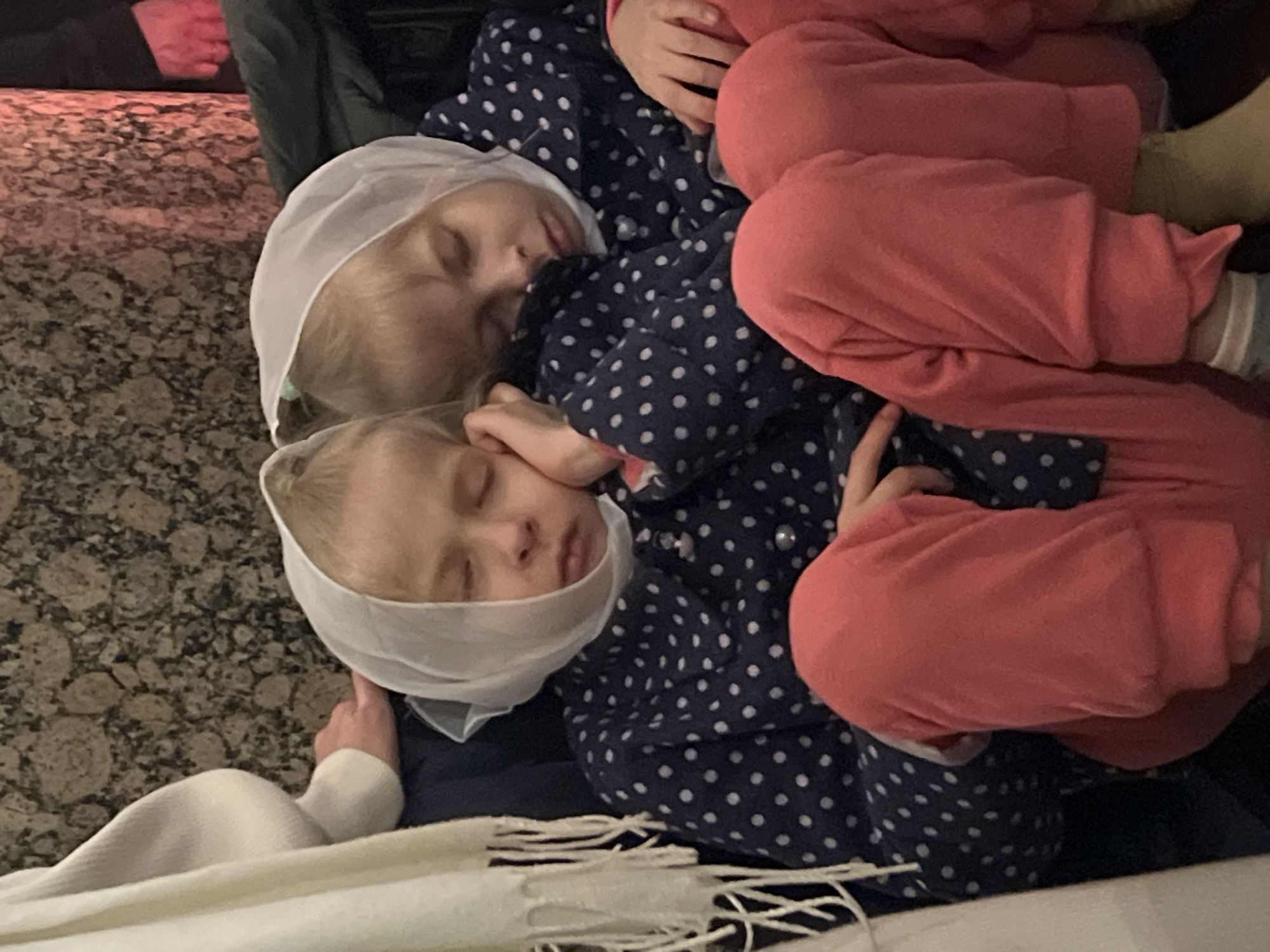 Two young children are sleeping on a surface covered with a blanket and a patterned shirt. Both children wear white head coverings. One child is dressed in a dark blue shirt with white polka dots, and the other is wearing pink clothing. The background includes a textured surface suggestive of flooring and the presence of other individuals nearby. There is no concrete evidence of the location being Saint Petersburg, Russia.