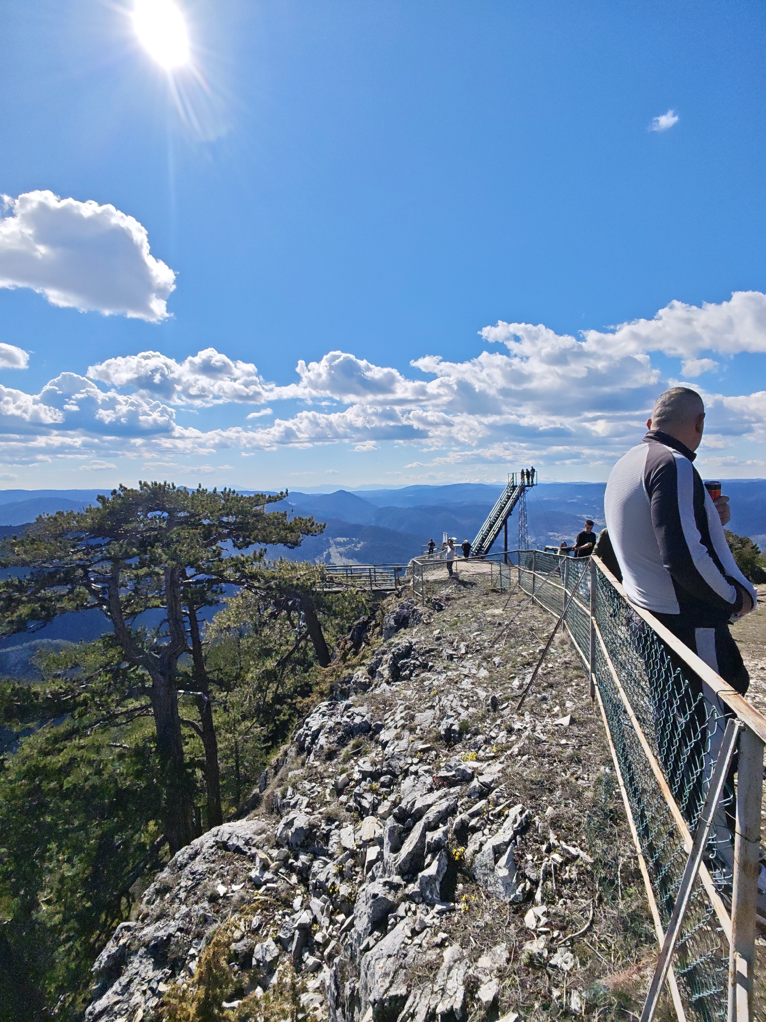 The image depicts a mountain overlook in Giovren, Bulgaria, featuring a man seated on a safety railing observing the landscape. A metal mesh fence lines the edge of the rocky terrain. In the background, a metal observation structure is visible. Mountains and valleys are spread out in the distance under a partially cloudy blue sky. The sun is prominent in the upper left corner. To the left, pine trees grow on the rocky slope. Other people are visible in the background near the observation structure.