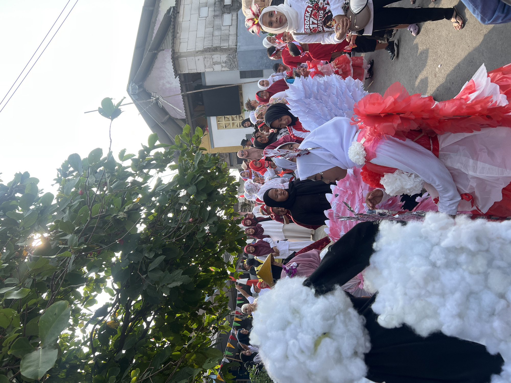 In Klapanunggal, Indonesia, a group of children participates in a parade celebrating Independence Day, marked by the "17 Agustus" inscription on a t-shirt. The procession is characterized by vibrant costumes, including white and red feathered wings on some children, symbolizing the Indonesian flag colors. Many participants are adorned in traditional Islamic attire, such as hijabs, with some girls wearing white outfits complemented by floral embellishments in red and white. A prominent figure in the foreground wears a large, white fluffy costume that obscures their lower body, possibly representing a cloud or a character. The scene is set outdoors, with overhead trees casting dappled sunlight, and residential buildings lining the street, indicating a community gathering. The overall atmosphere is festive and celebratory, with onlookers gathered along the parade route.
